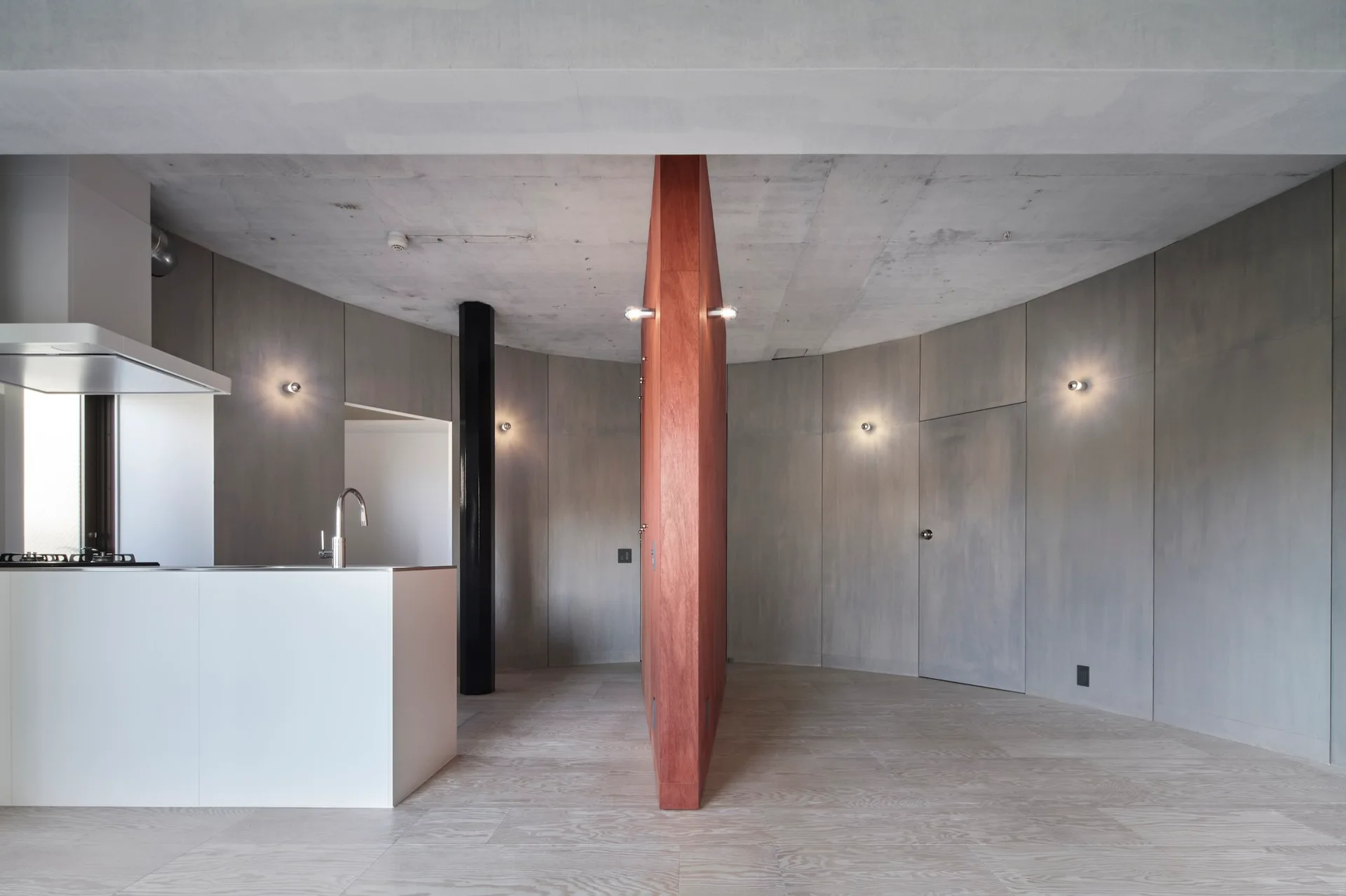 A modern minimalist interior featuring curved gray walls, a central red wood partition, and a white kitchen island beneath an exposed concrete ceiling.