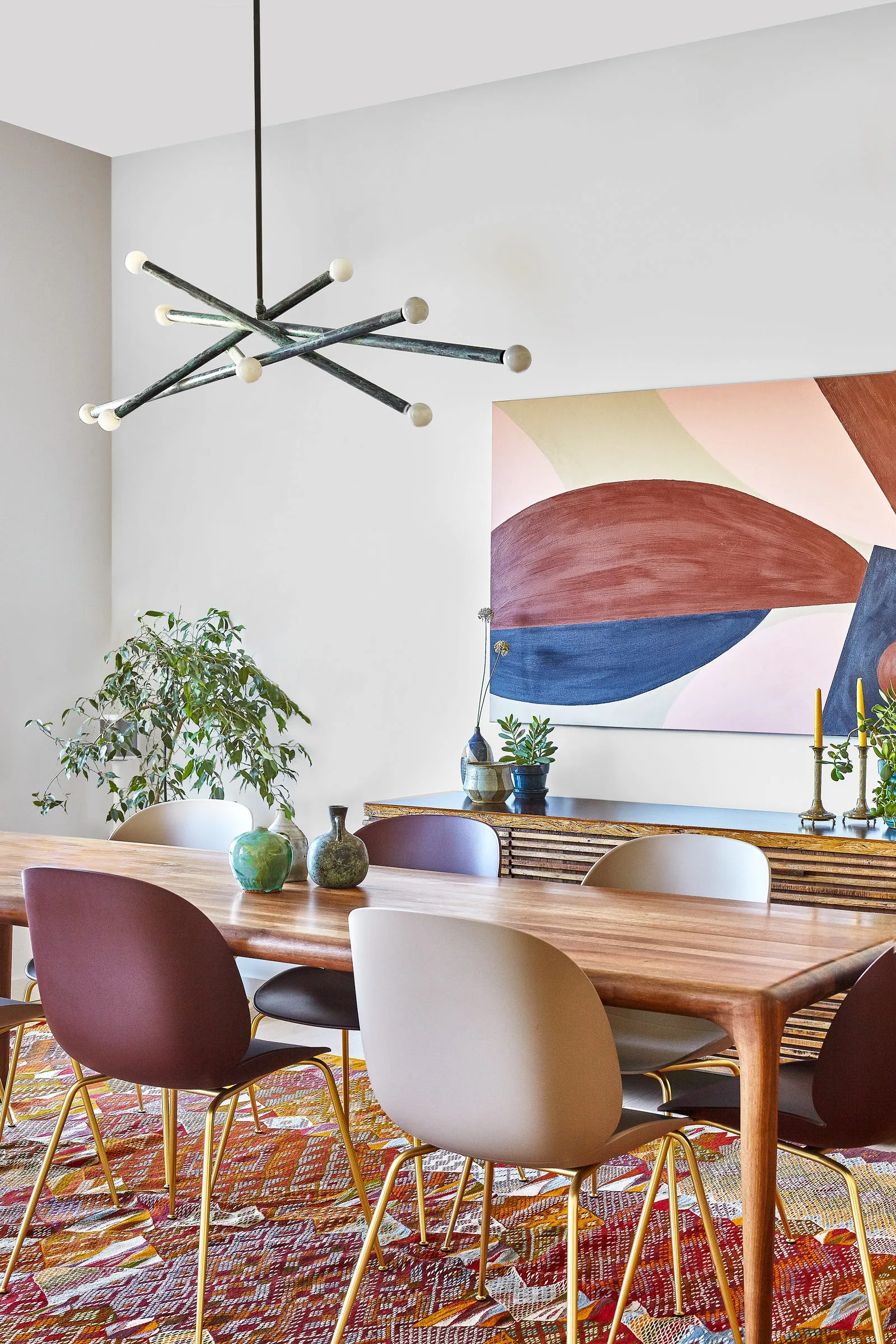 A modern dining room features a wooden table, chairs in mismatched neutral tones, a colorful geometric rug, and a large abstract painting.