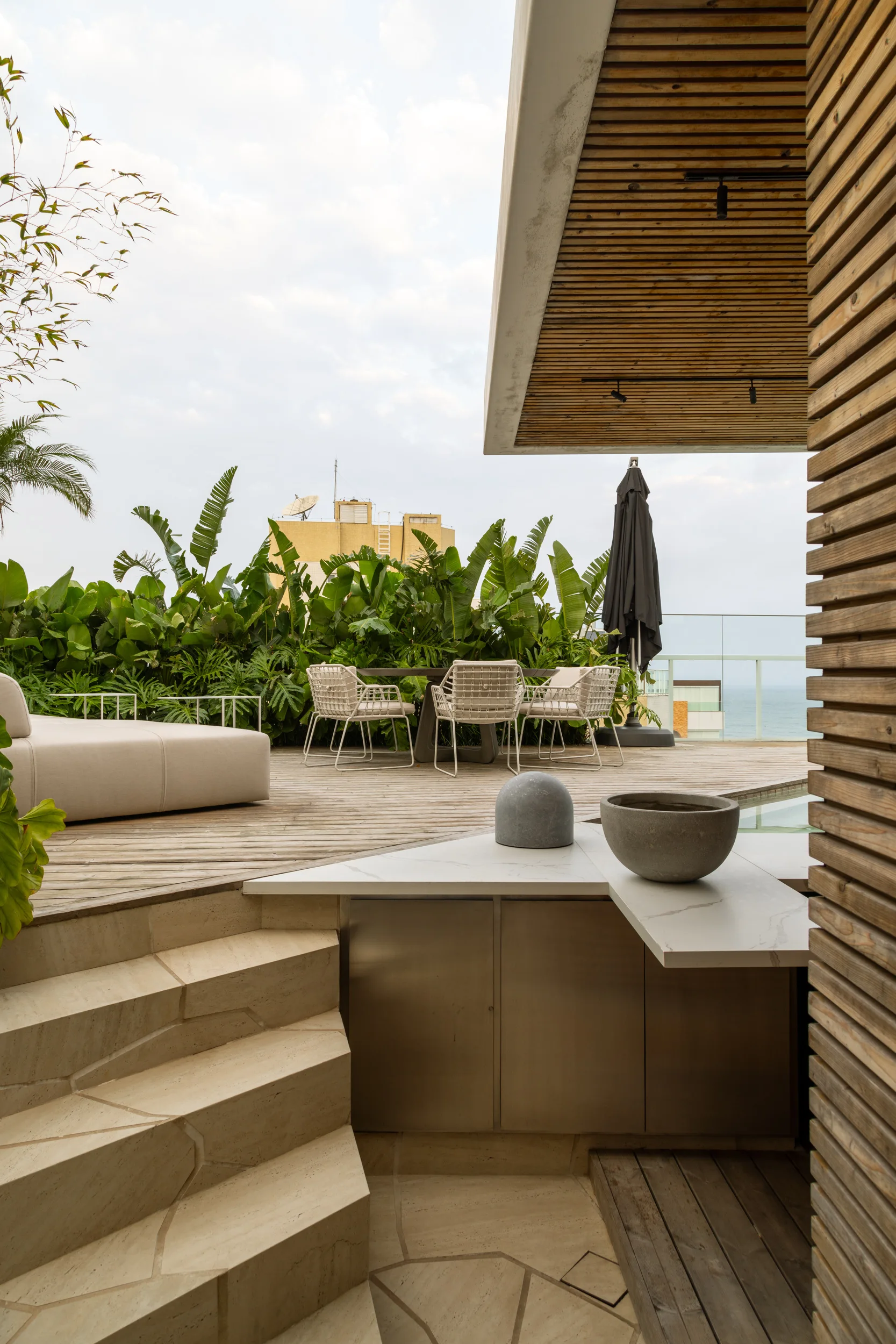 A modern wood terrace features a dining set, curved stone steps leading to built-in cabinetry, and a slatted wood overhang against tropical greenery.
