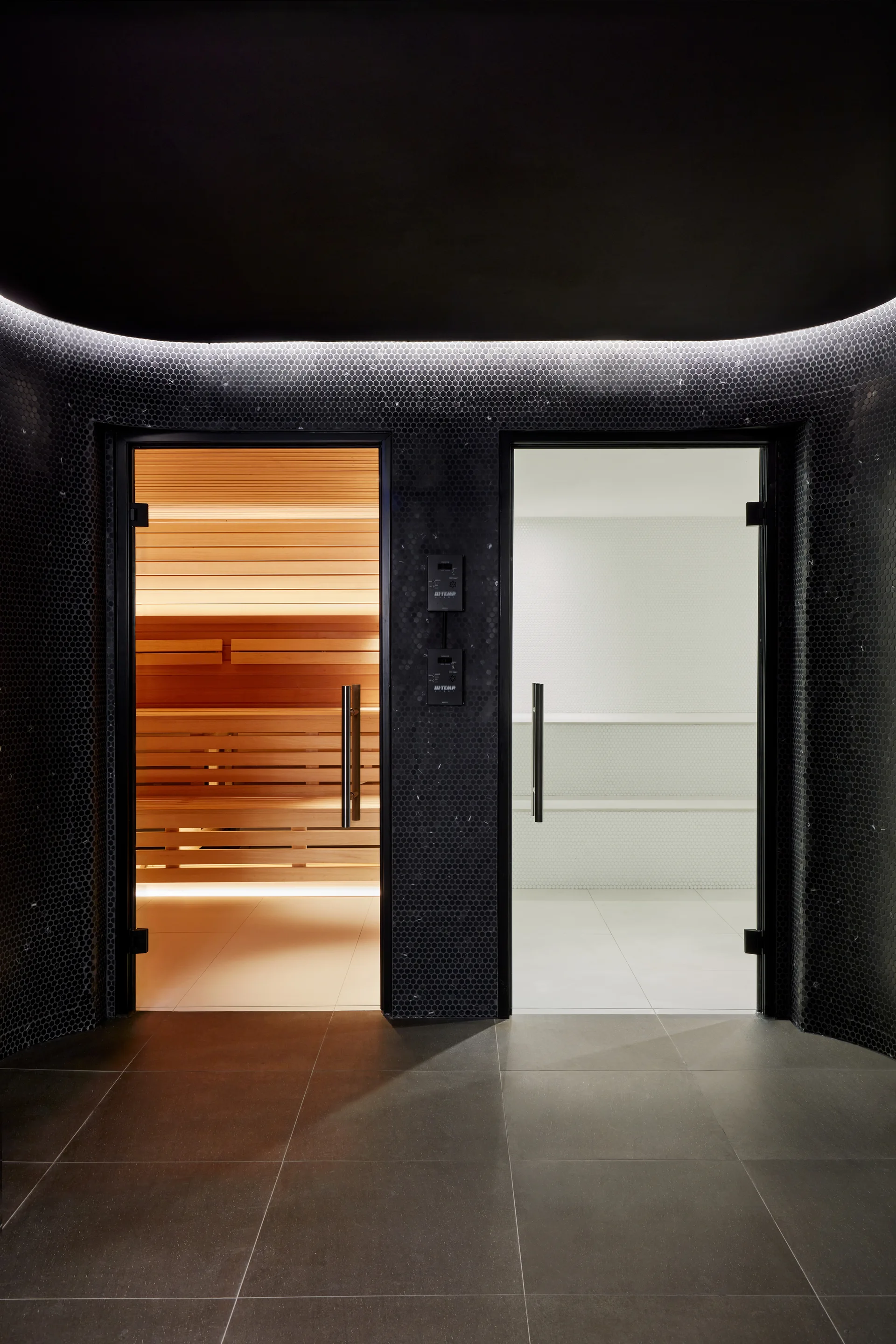 Two glass doors set in a curved black penny tile wall reveal a warm wood-slat sauna and a bright white tiled steam room.