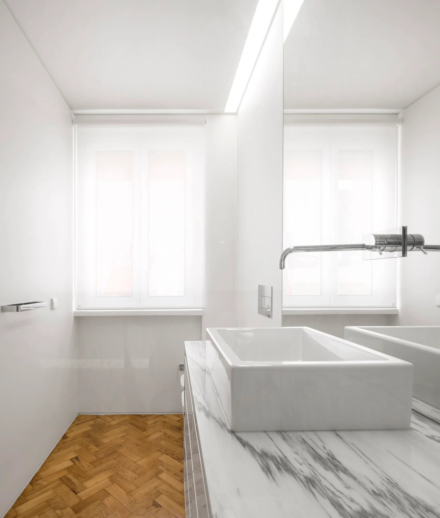 A minimalist bathroom features a white vessel sink on a veined marble vanity next to a herringbone patterned wood floor.