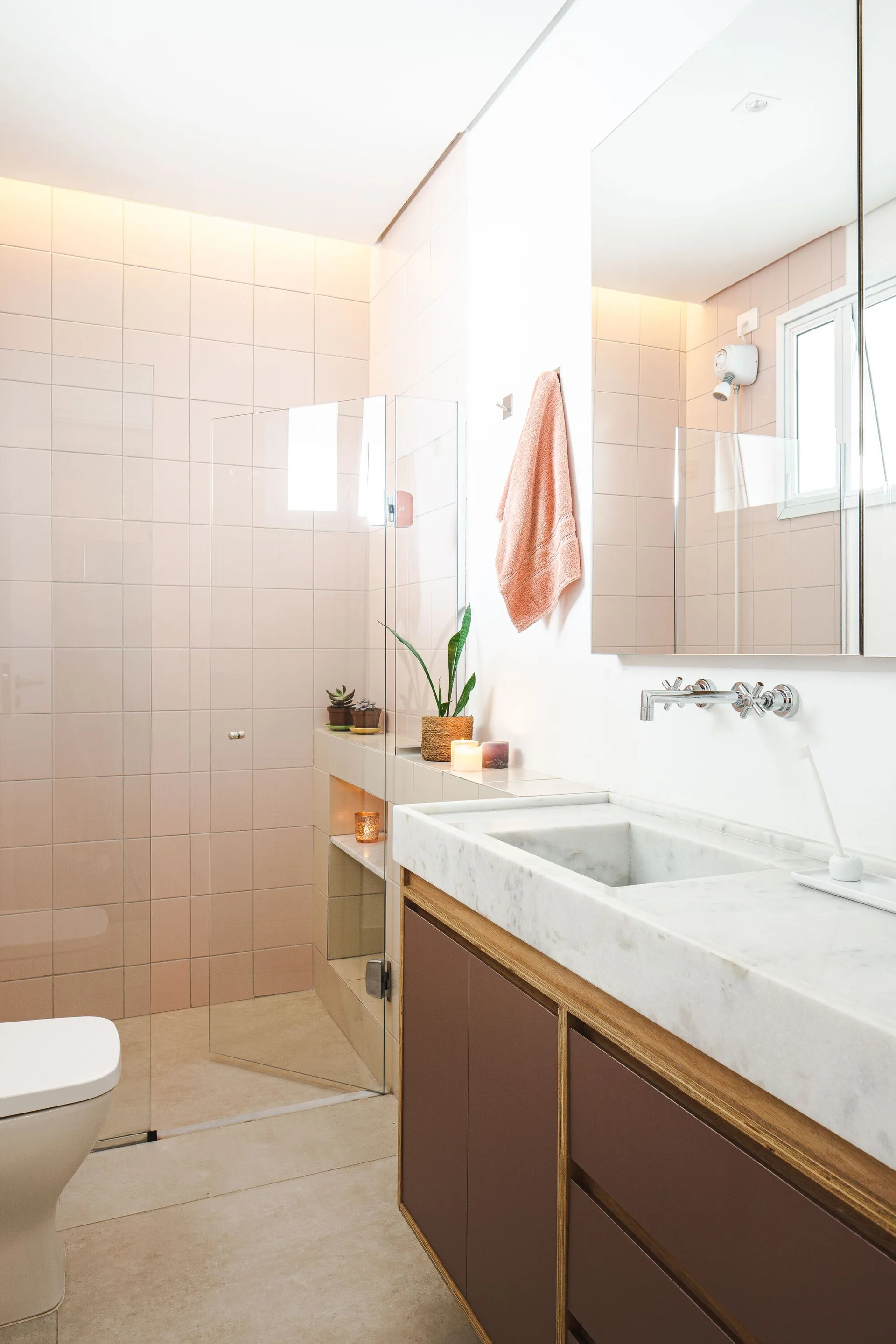 A modern bathroom featuring pale pink wall tiles, a marble vanity with brown cabinets, a chrome faucet, and a glass shower door.