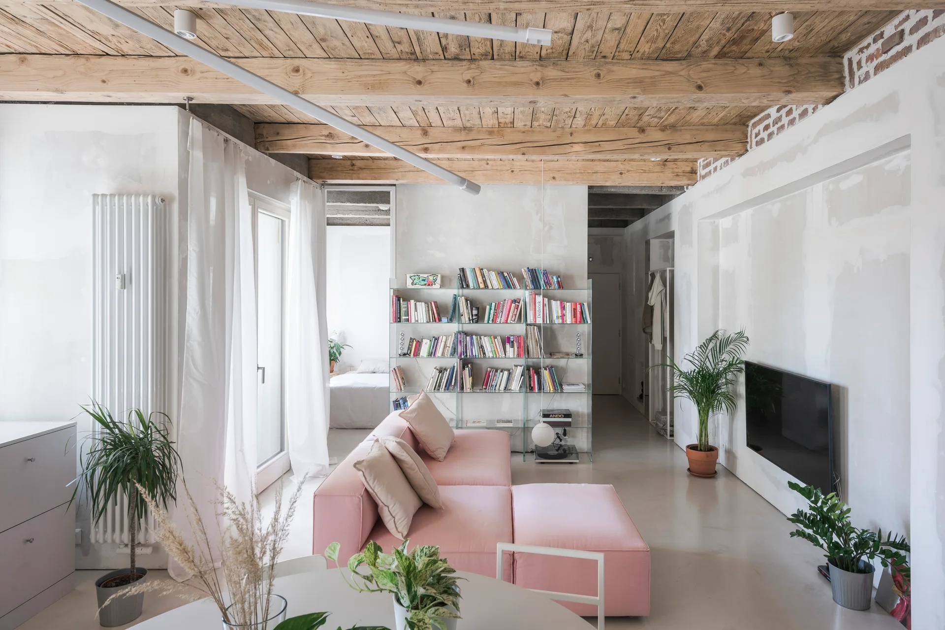 A minimalist living room features a low pink modular sofa, a clear glass bookcase, raw plaster walls, and exposed wooden ceiling beams.
