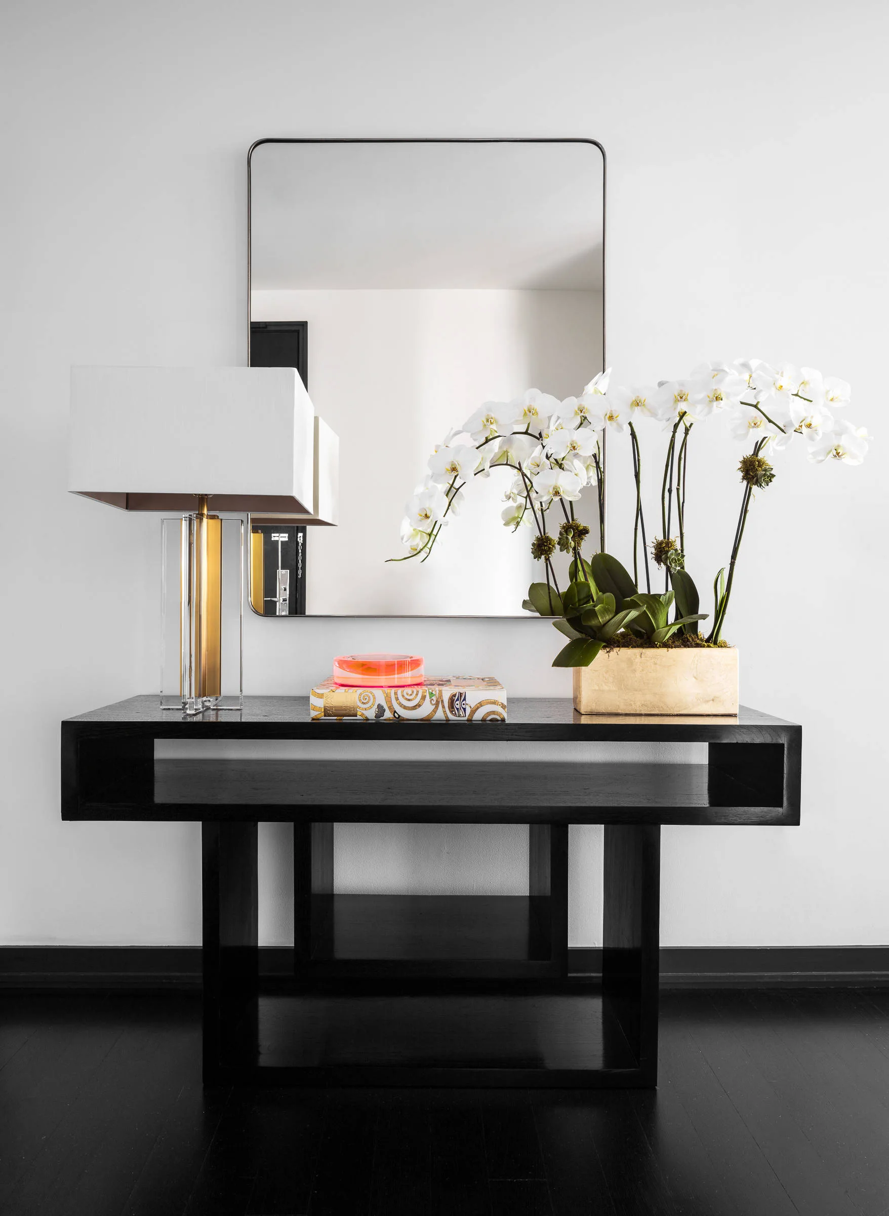 A black wooden console table displays a tall glass table lamp, a patterned book with a coral dish, and white orchids beneath a rectangular mirror.