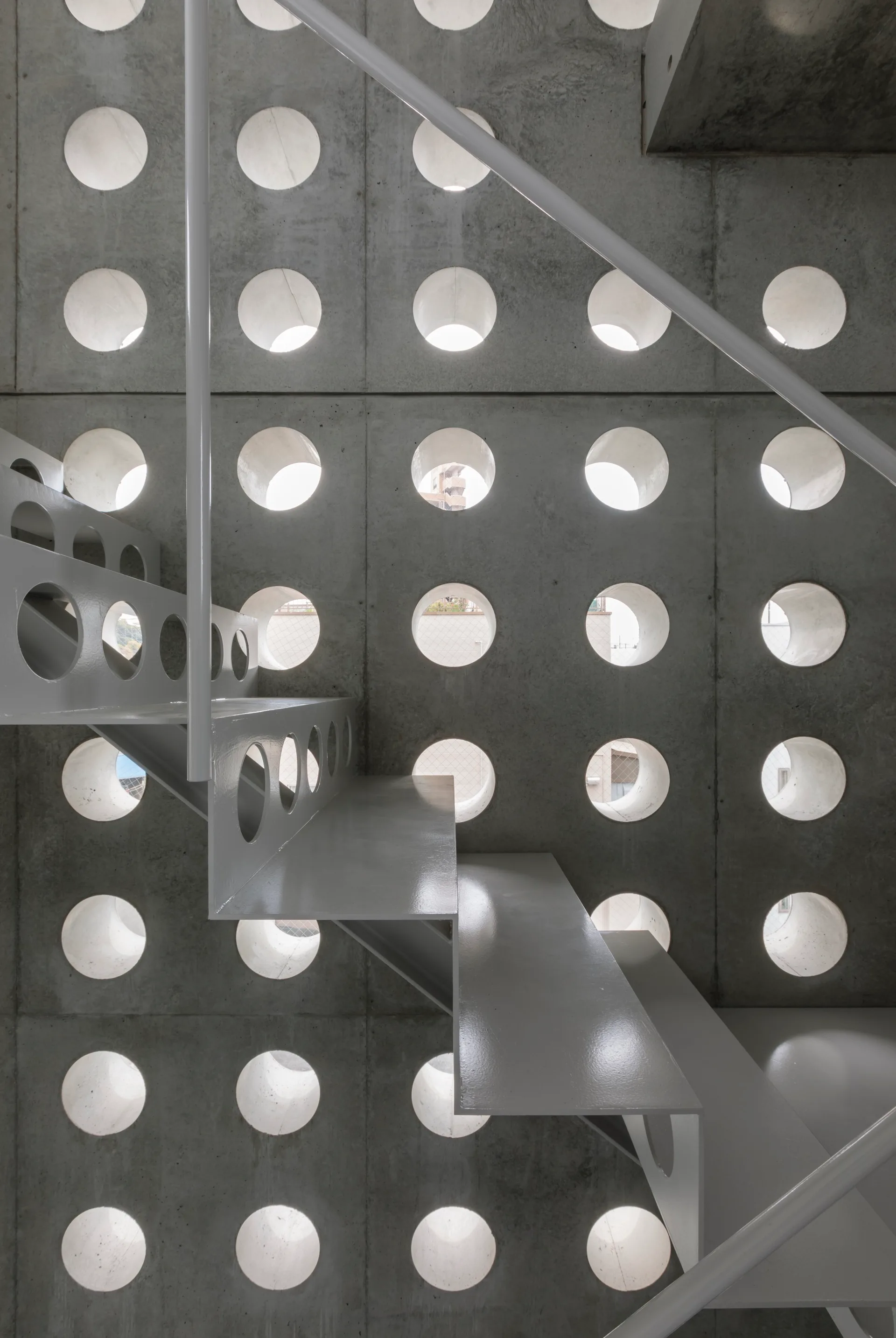 A glossy white metal staircase stands in front of a raw grey concrete wall patterned with a grid of large circular openings.