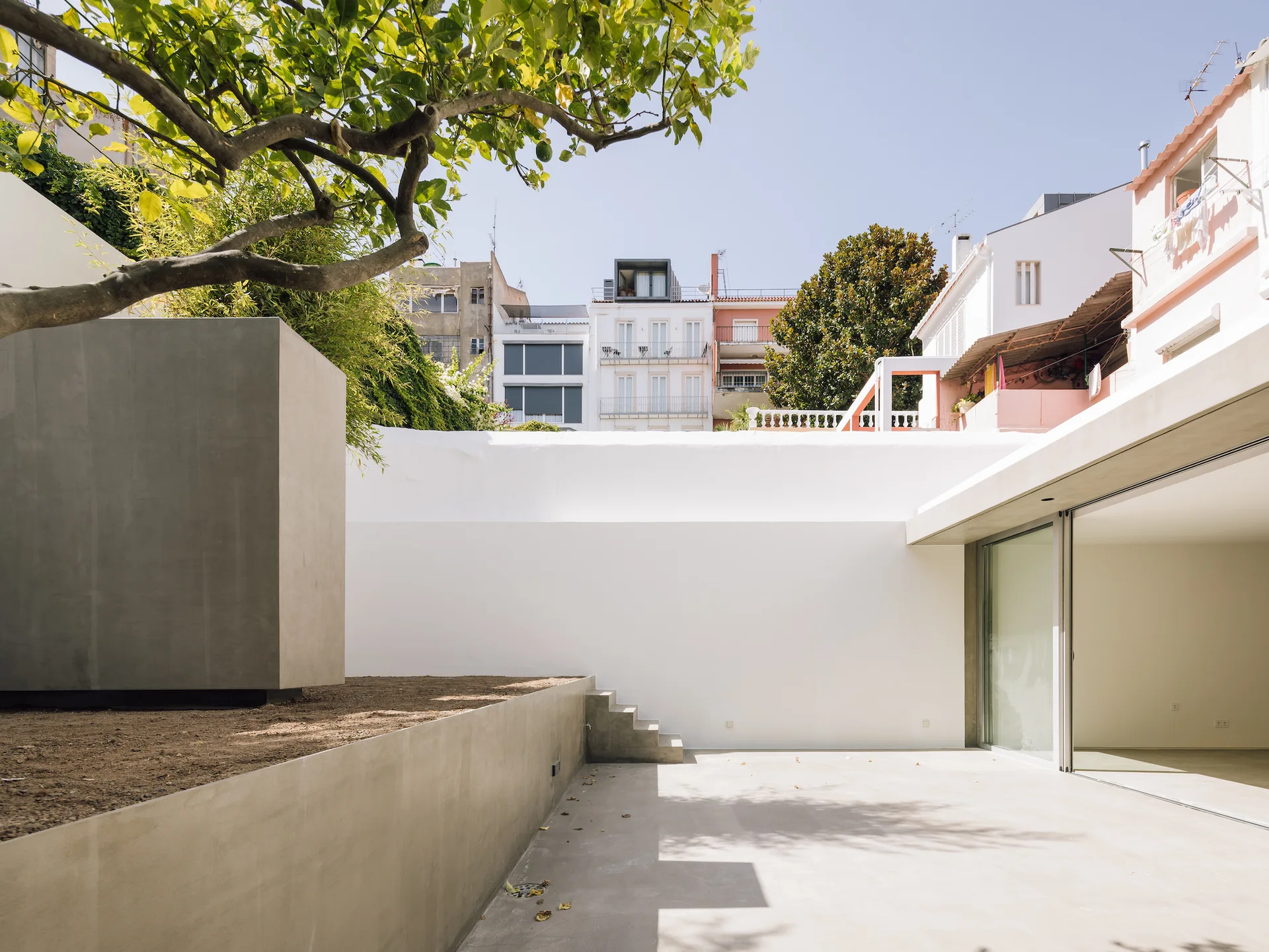 A minimalist urban patio features a smooth concrete floor, a raised concrete planter with soil and a tree, and a high white perimeter wall.