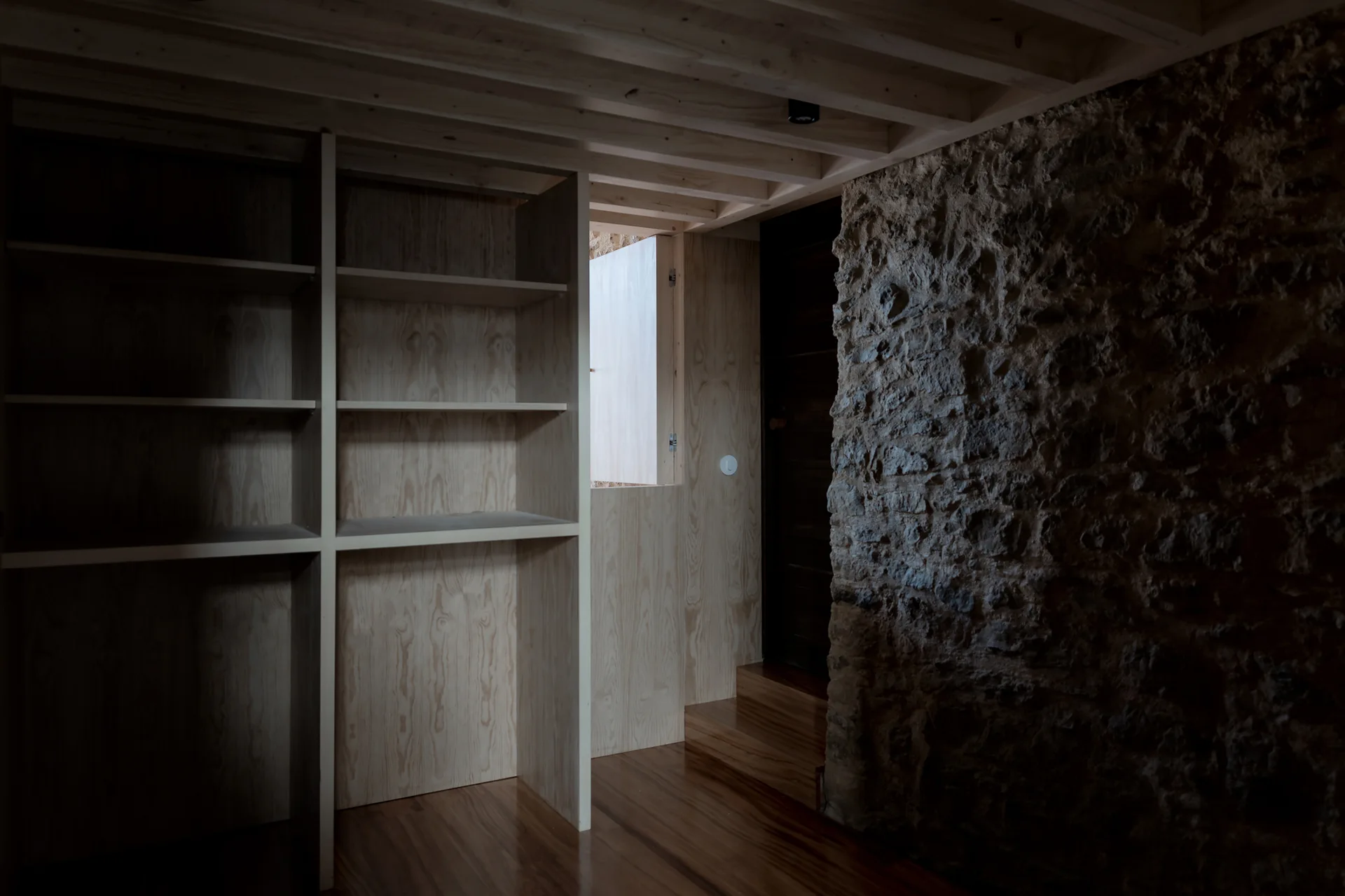 Built-in plywood shelving and an exposed beam ceiling stand beside a rough natural stone wall in a dimly lit rustic interior.