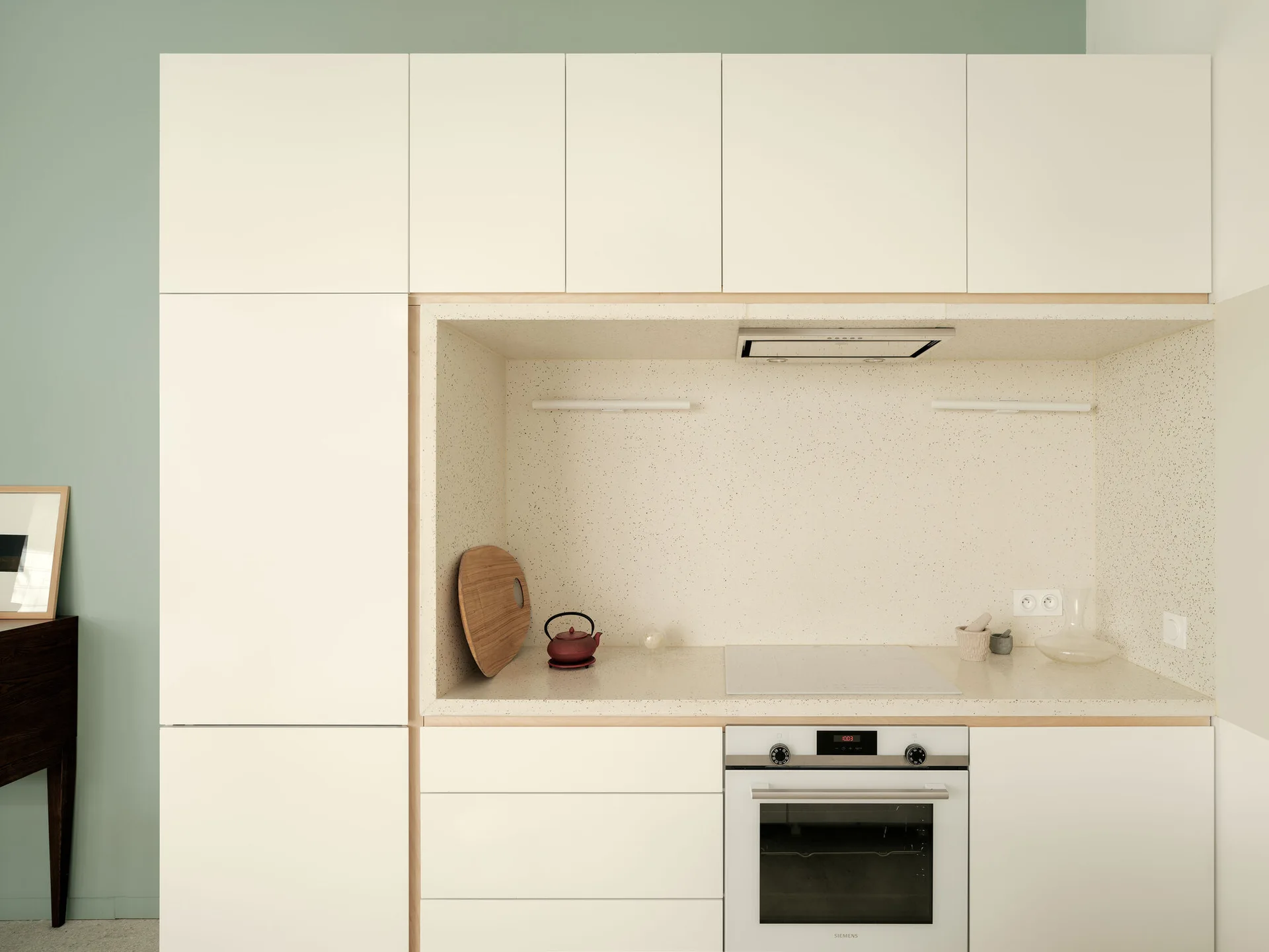 Minimalist white handleless kitchen cabinets feature a recessed cooking niche lined with speckled beige stone and framed in light wood trim.