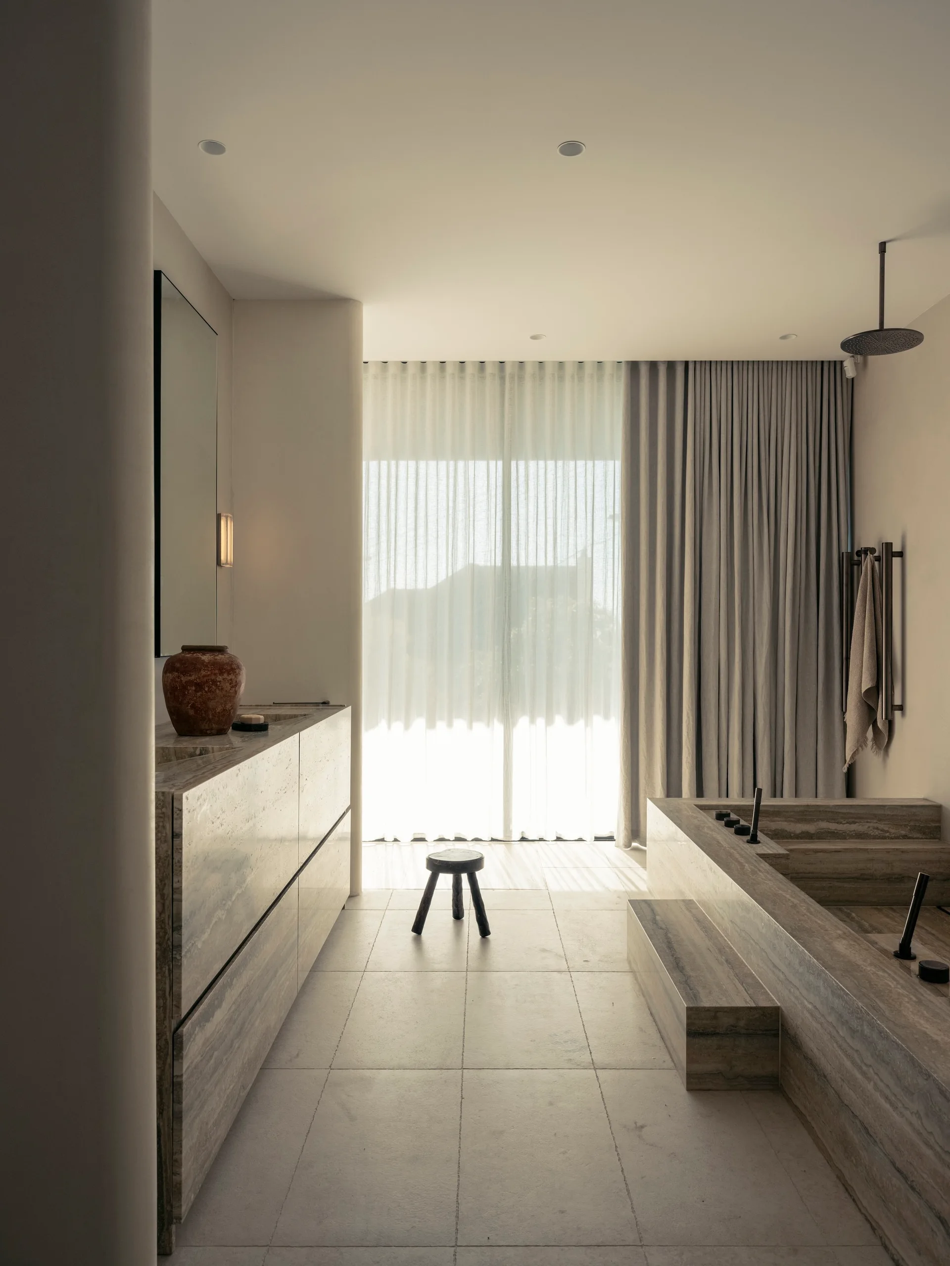 A minimalist beige bathroom features a travertine-clad vanity and built-in stone bathtub, with sheer floor-to-ceiling drapes filtering natural light.