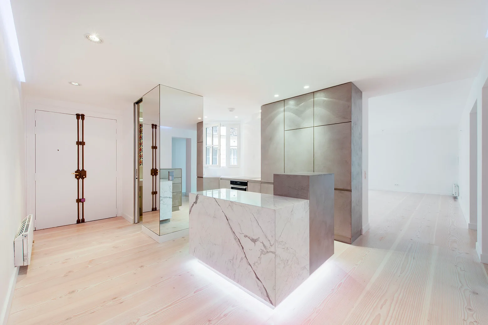 A minimalist modern kitchen features a marble island with base lighting, grey cabinetry, a mirrored column, and light wood plank flooring.
