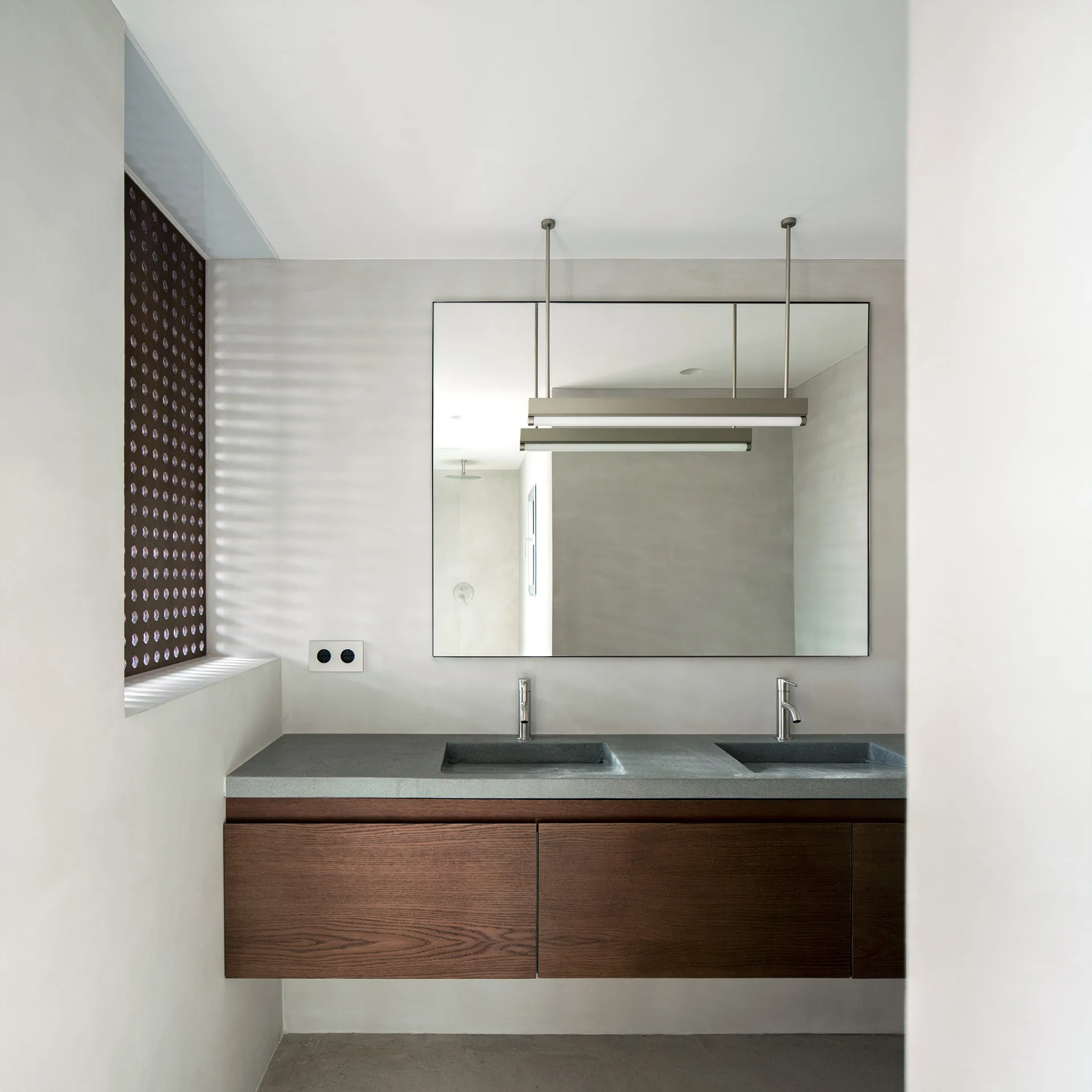 A modern bathroom with a dark wood floating vanity, grey stone-like countertop with two integrated sinks, a large mirror, and a hanging linear light.