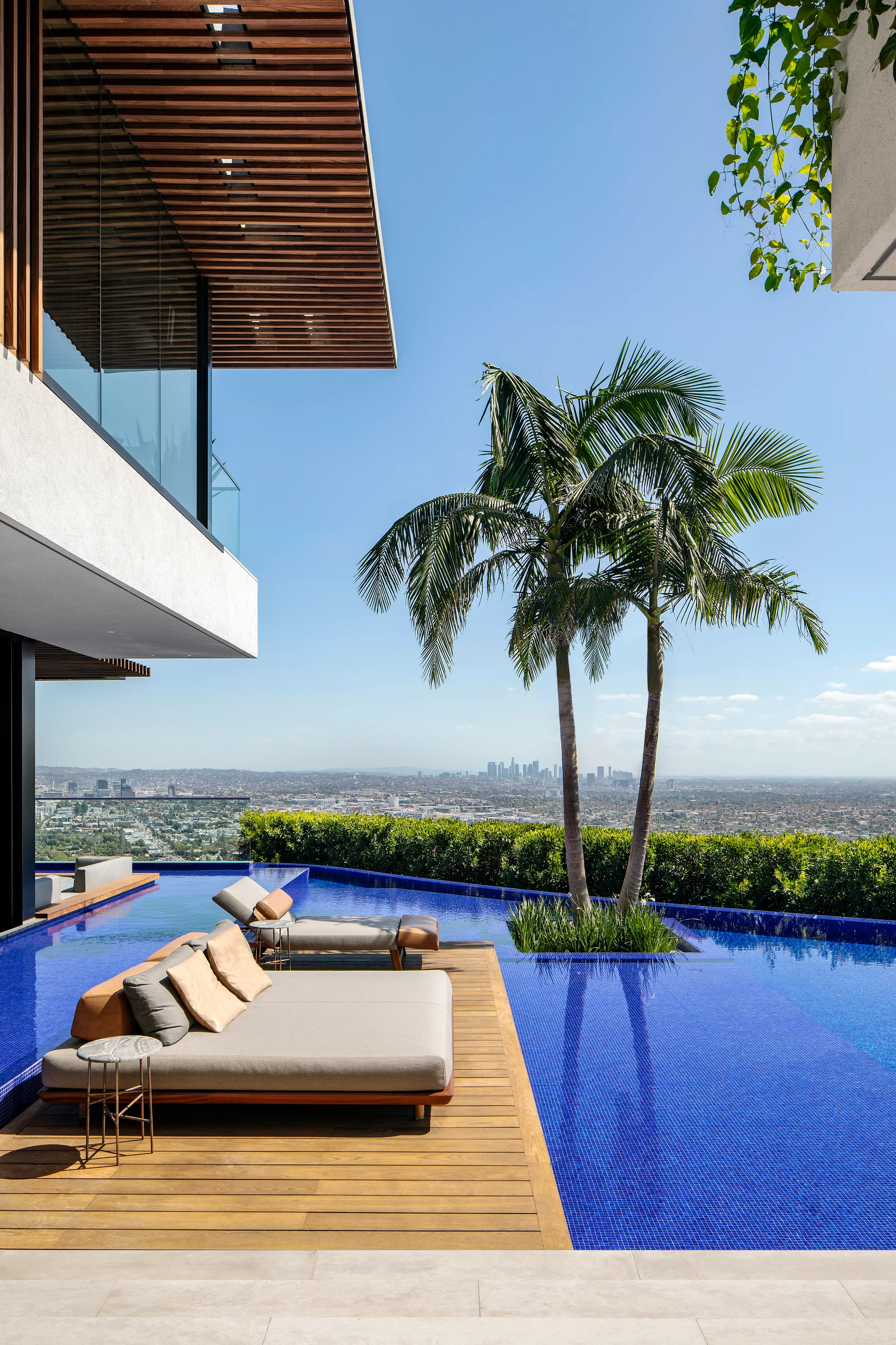 An outdoor infinity pool with blue tiles, a wooden deck platform holding two loungers, a palm tree on a small island, and a city skyline background.