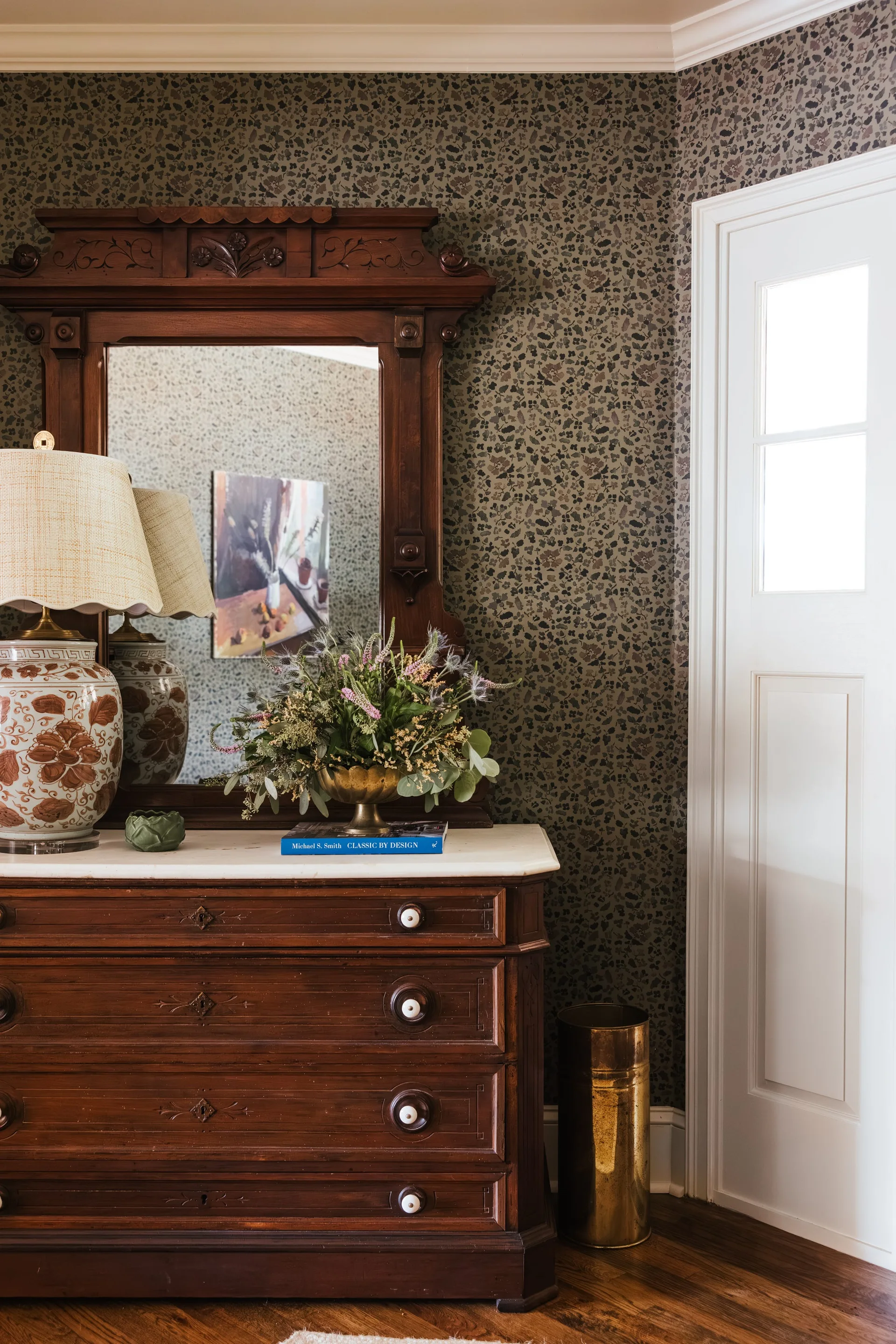 A vintage dark wood dresser with a white marble top is positioned against dark floral wallpaper next to a white paneled door.
