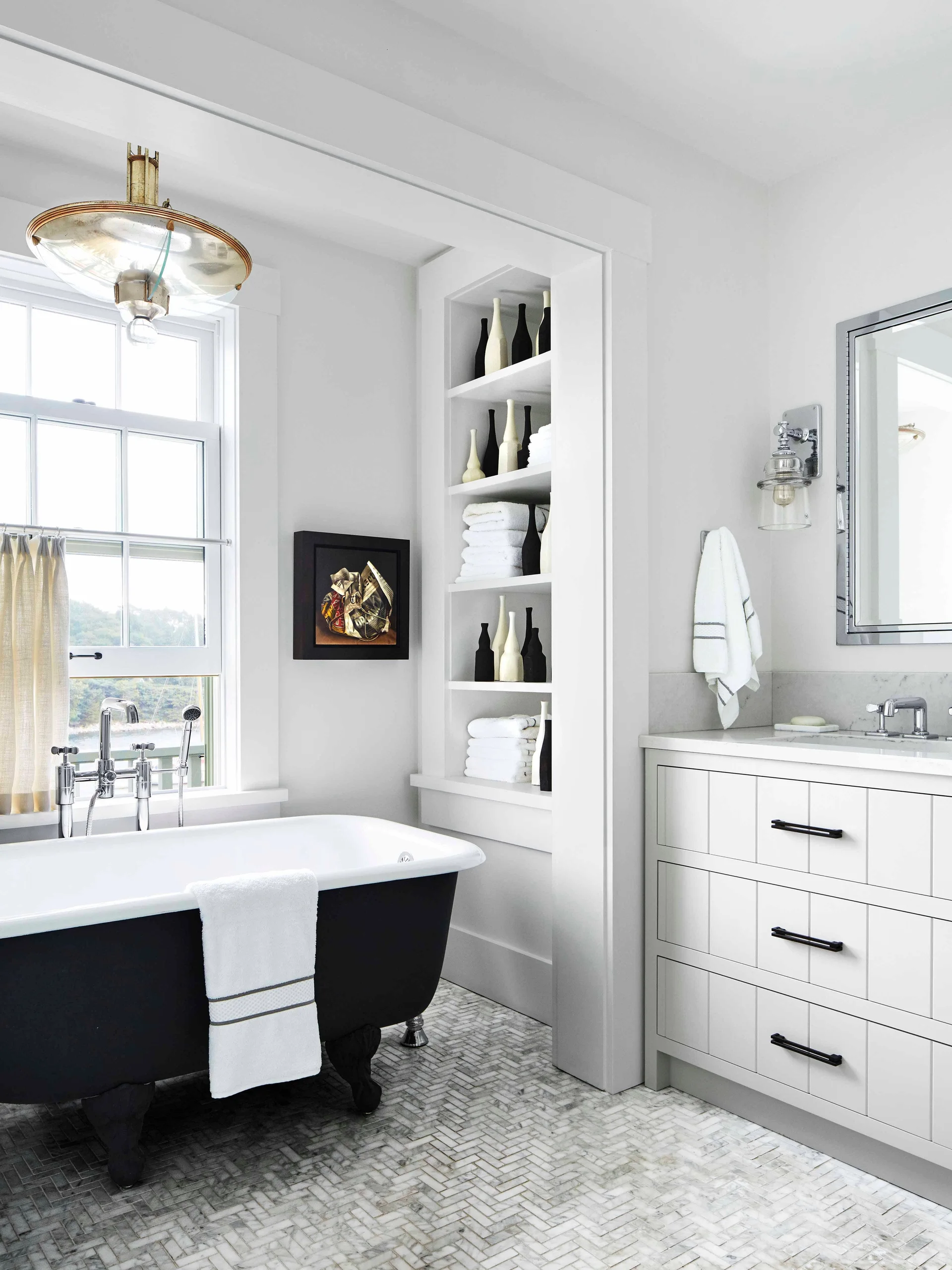 A bathroom features a black-bottom clawfoot bathtub next to a window, a white vanity with black drawer pulls, and a herringbone marble tile floor.