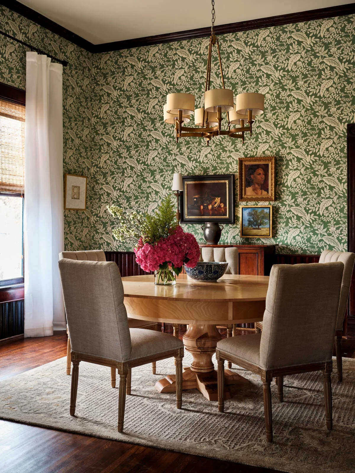 A traditional dining room featuring a round wooden pedestal table, upholstered chairs, green patterned wallpaper, and a gold chandelier.