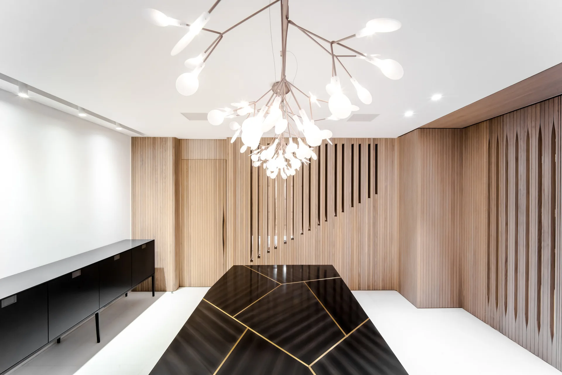 A contemporary dining room features vertical wood slat walls, a black geometric dining table with gold inlay, and a branching white chandelier.
