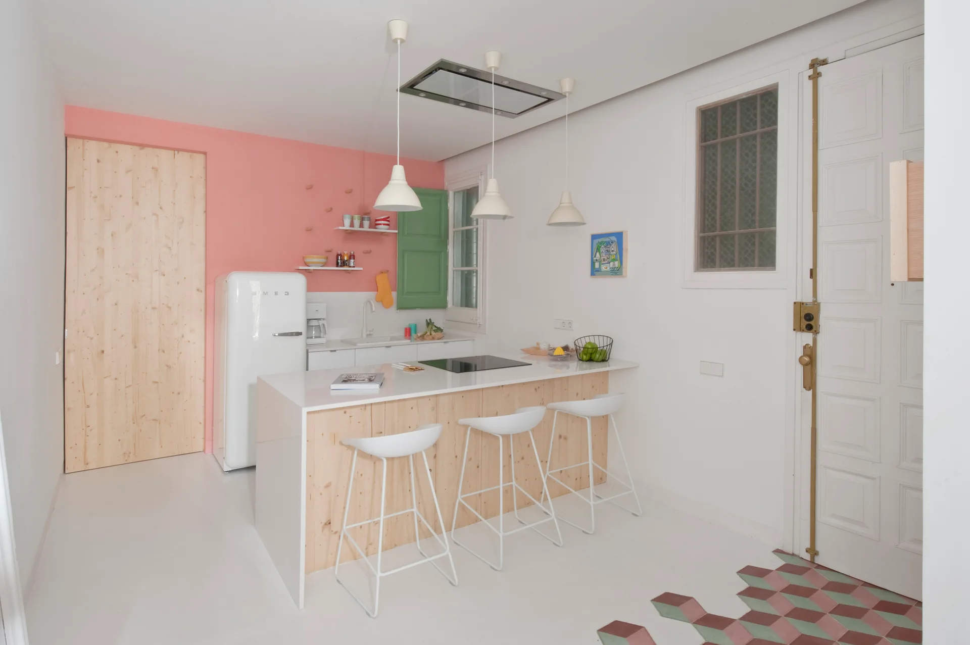 A modern white kitchen features a soft pink accent wall behind a white retro refrigerator and a light wood island with white bar stools.