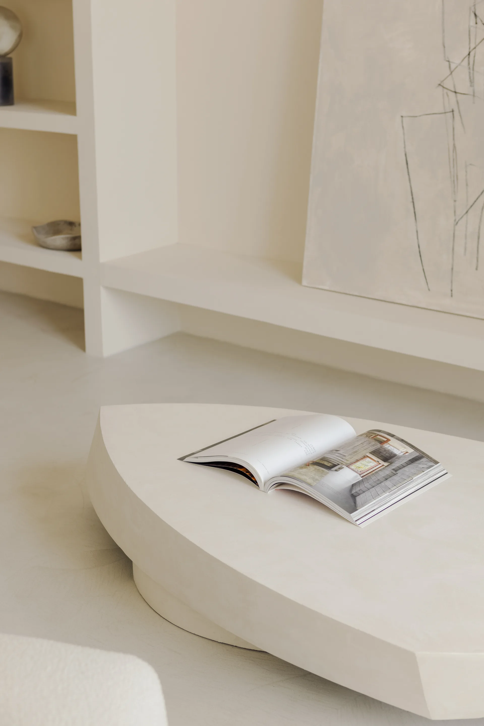 A low organic-shaped coffee table with an open book sits in front of built-in recessed plaster shelving displaying minimal decor.