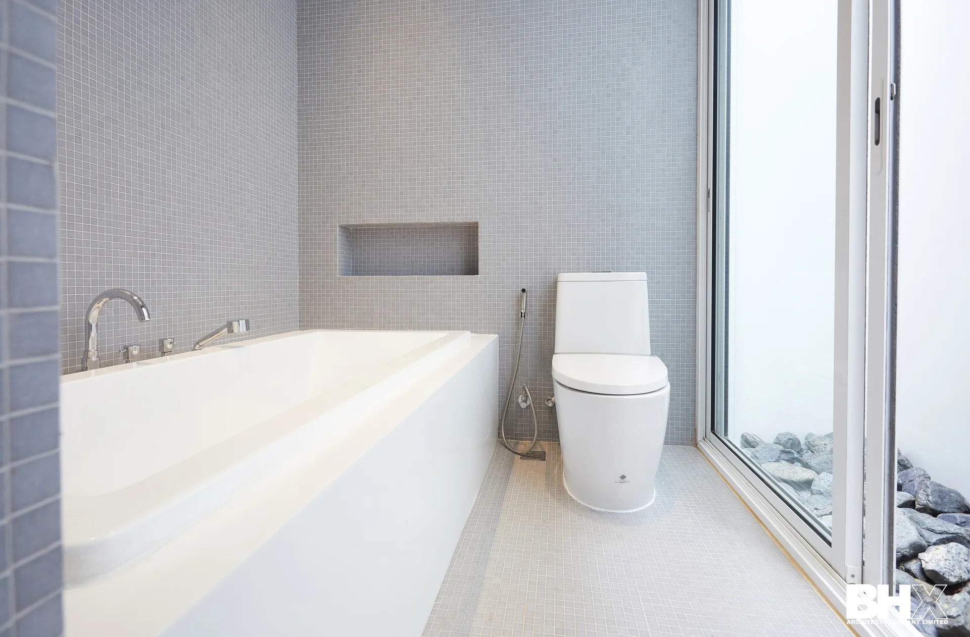A modern bathroom features grey square mosaic tiled walls, a white built-in bathtub, a toilet, and a glass sliding door looking out to stones.