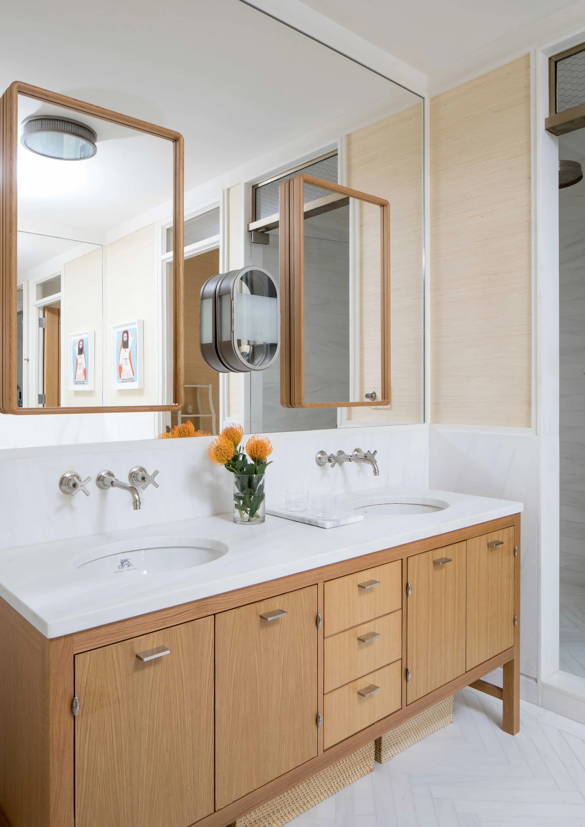 A light wood double bathroom vanity features white marble countertops, wall-mounted faucets, twin wood-framed mirrors, and textured beige wallpaper.