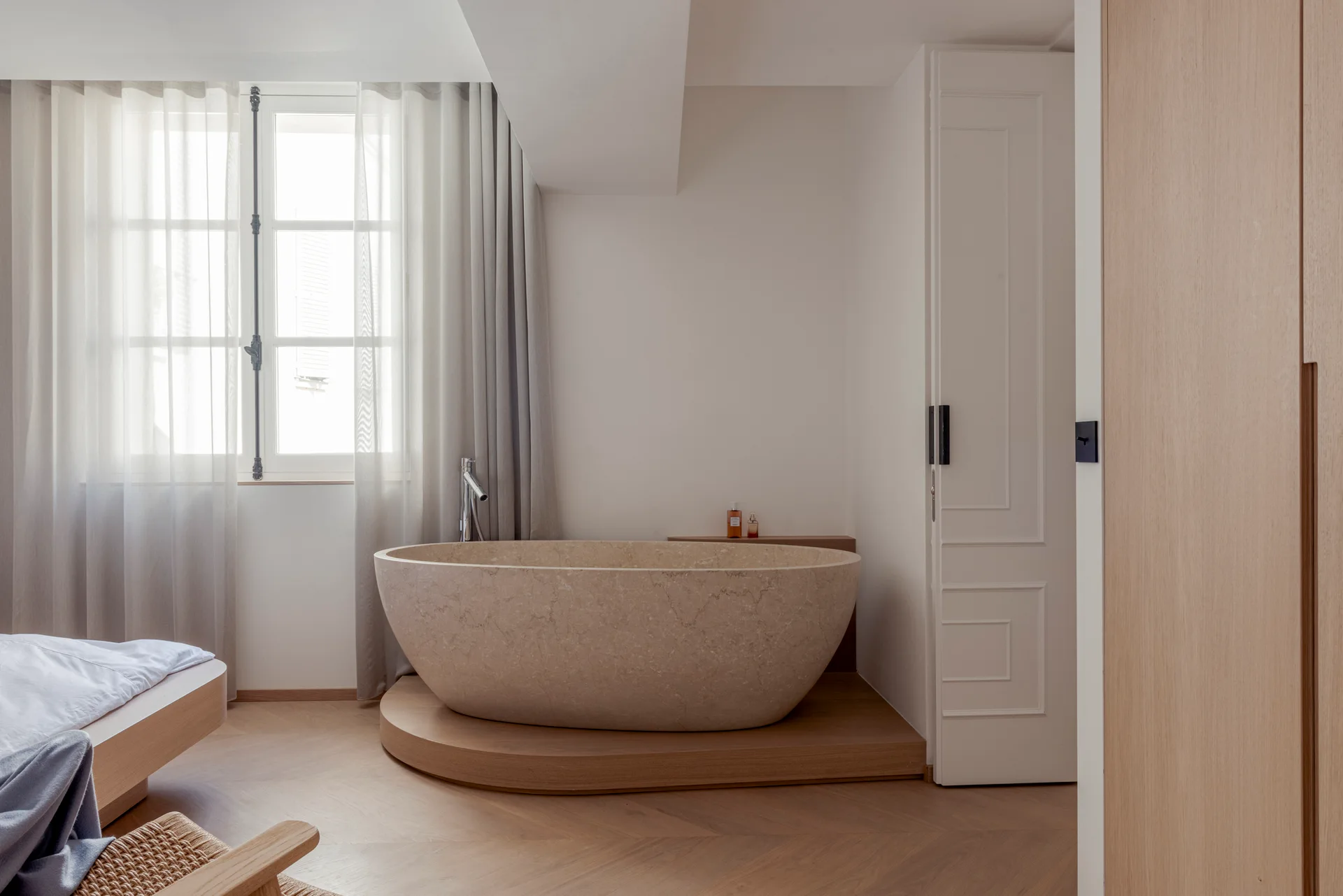 A beige stone freestanding bathtub sits on a raised wooden platform next to a window with sheer curtains and light wood herringbone flooring.