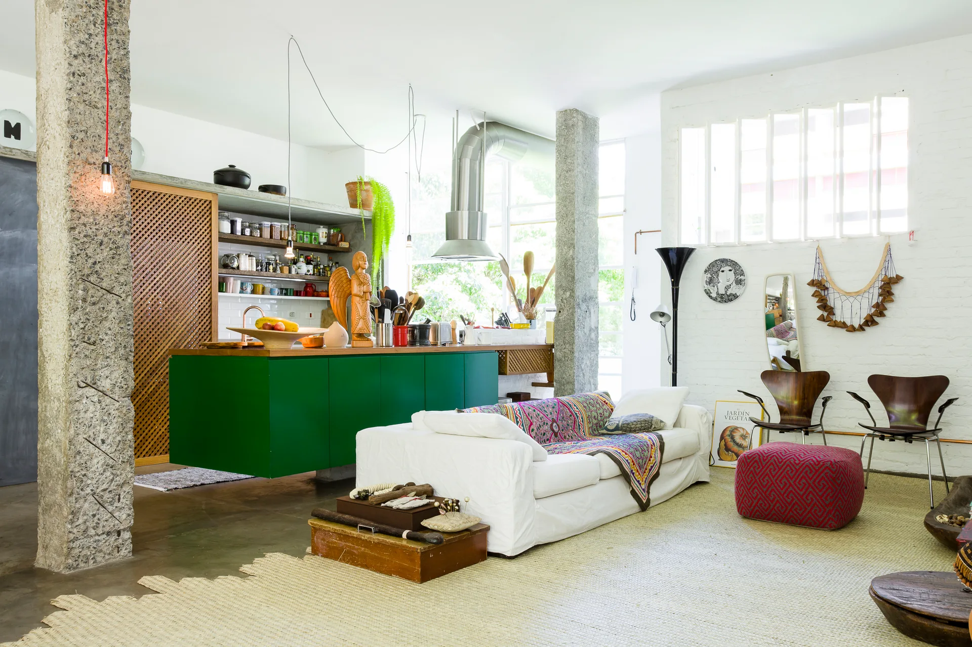 A bright open-plan loft featuring a white sofa on a woven rug, a vibrant green kitchen island, and textured stone pillars.