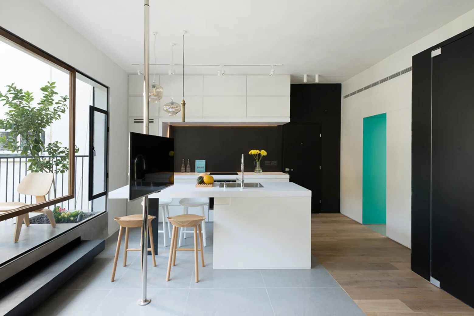 A minimalist kitchen features a white central island with wood stools, a pole-mounted TV, a black accent wall, and a bright teal doorway.