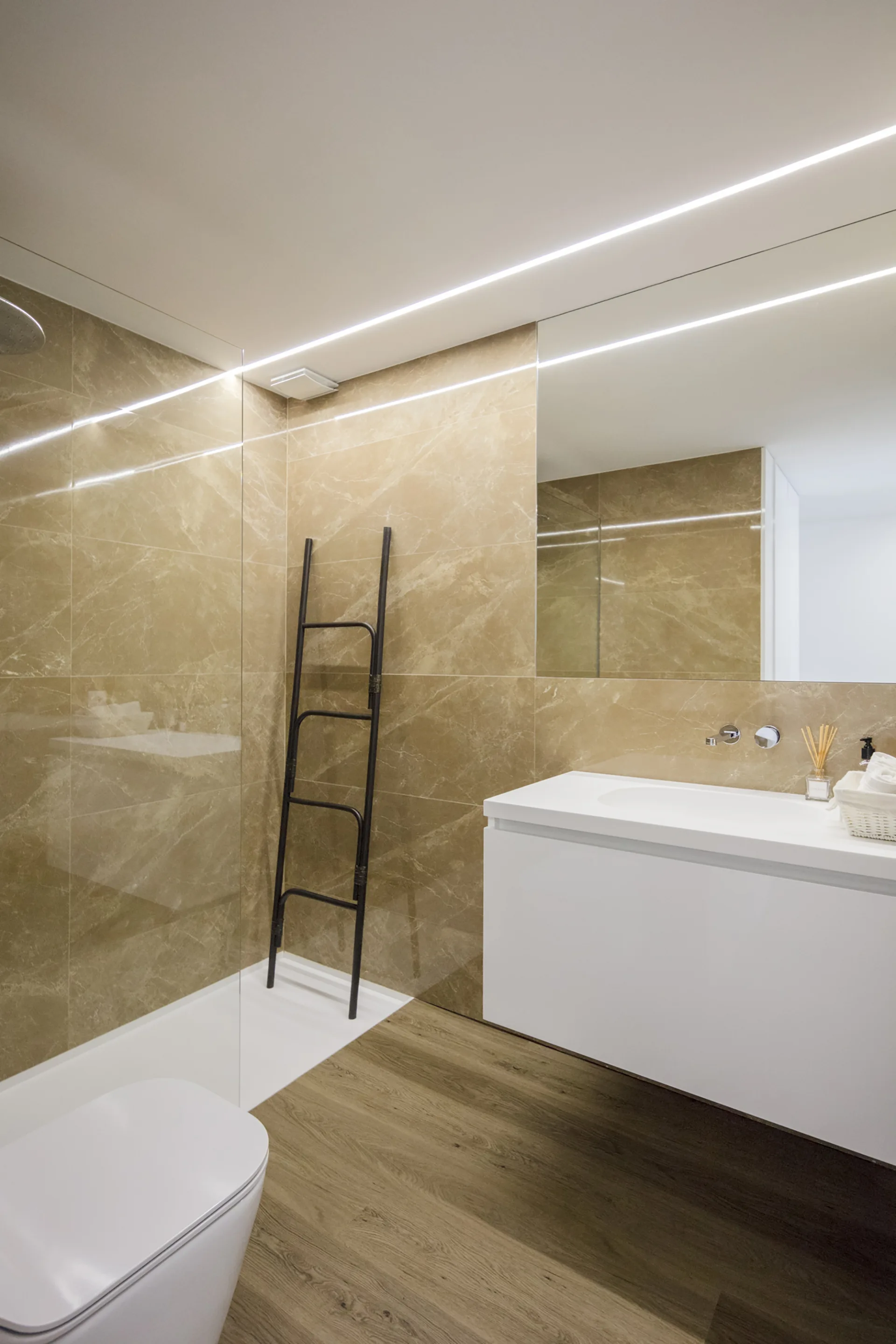 A modern bathroom with glossy beige marble wall tiles, a floating white vanity, a glass shower partition, and a black lean-to ladder.