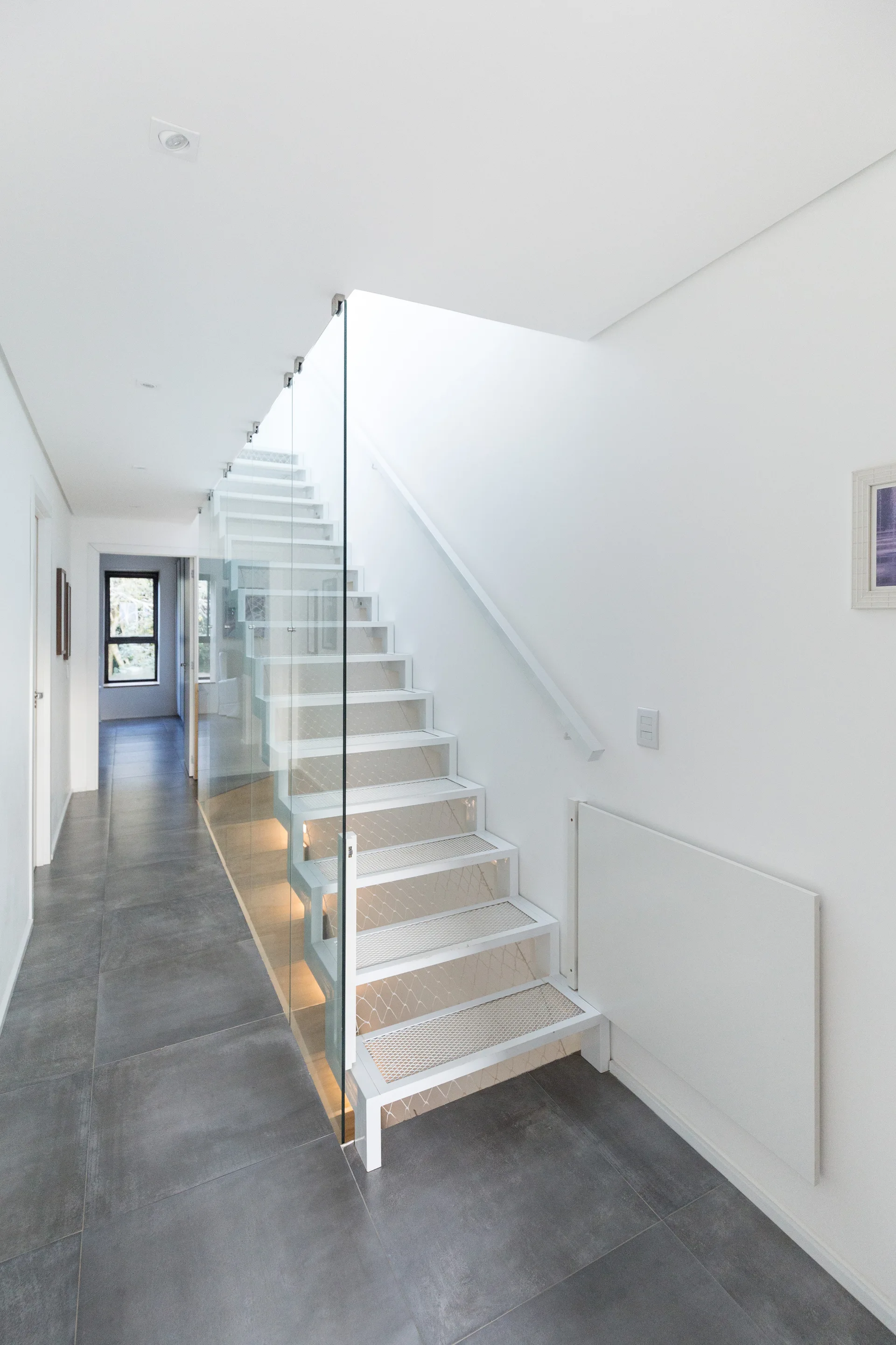 A modern white metal staircase with open mesh treads and a clear glass railing stands in a hallway with large grey floor tiles.