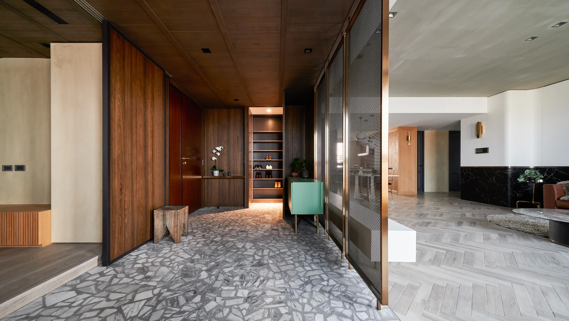 A modern entryway featuring dark wood wall paneling, an irregular marble mosaic floor, and a metal mesh room divider next to a green console.