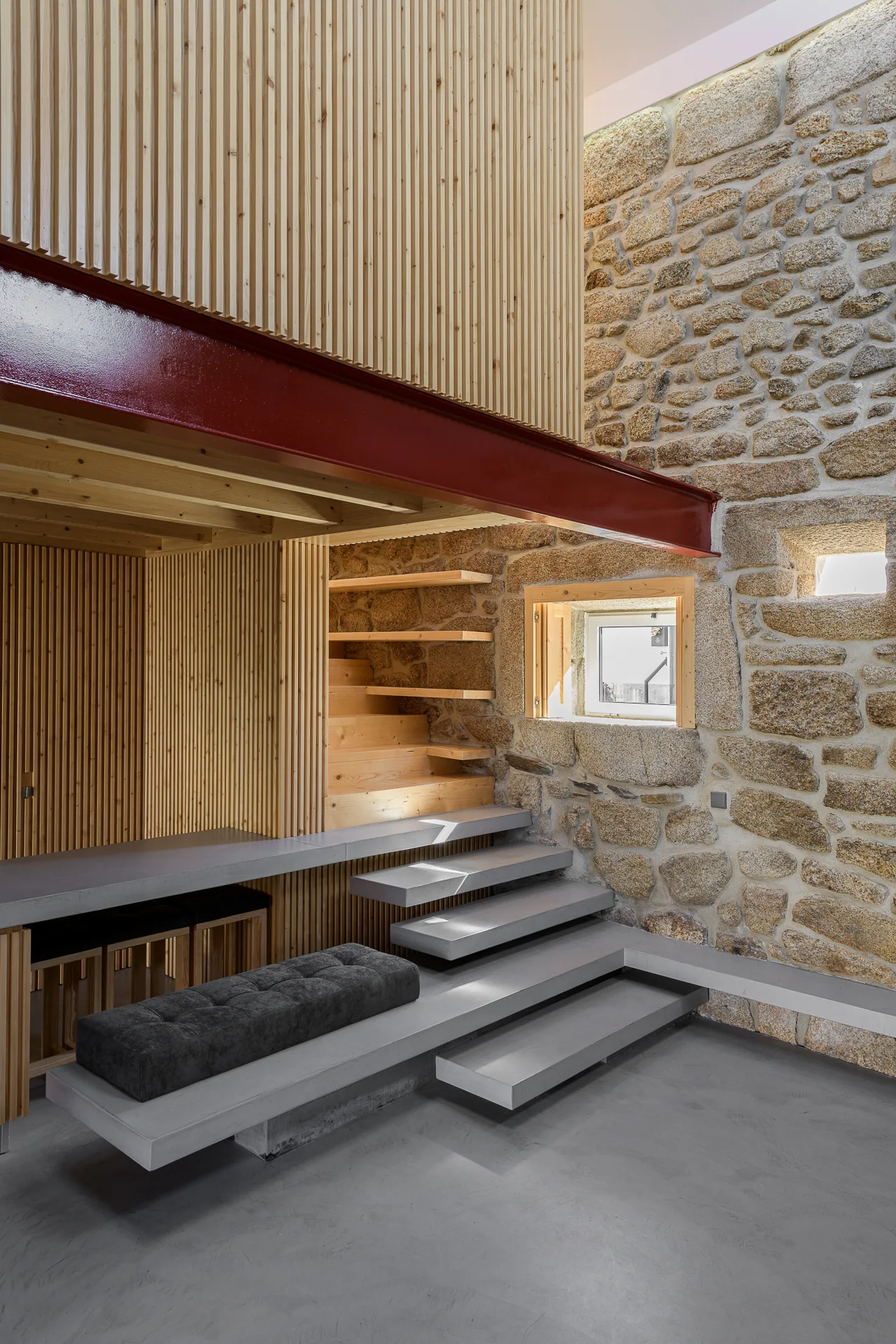 Minimalist floating concrete steps lead upward alongside a rough stone wall, balanced by vertical light wood slats and a dark red steel beam.