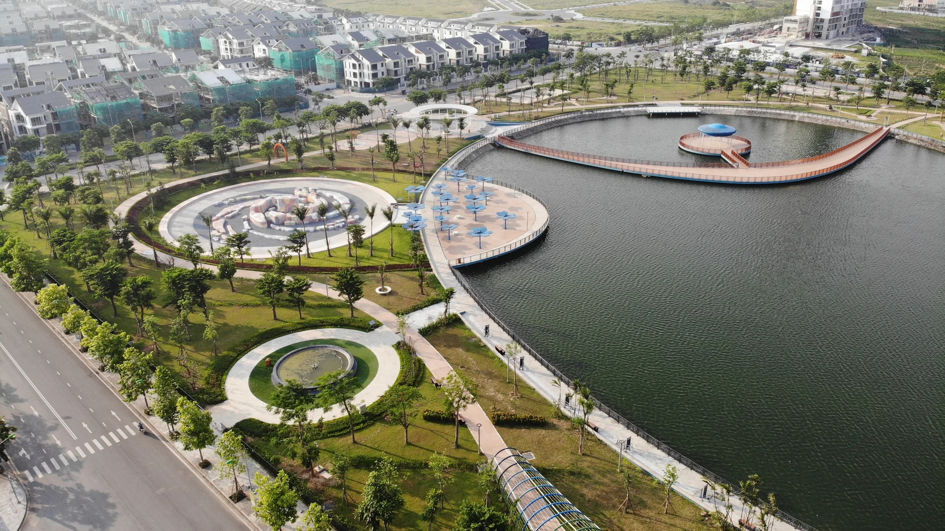 Aerial view of a landscaped modern park featuring a large lake, a curved pedestrian bridge, circular paved walkways, and green lawns.