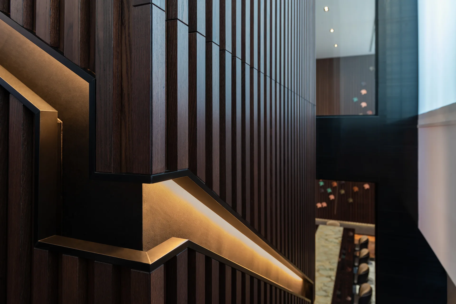 A close-up of a dark wood slatted staircase wall with an angular handrail illuminated by built-in warm lighting.