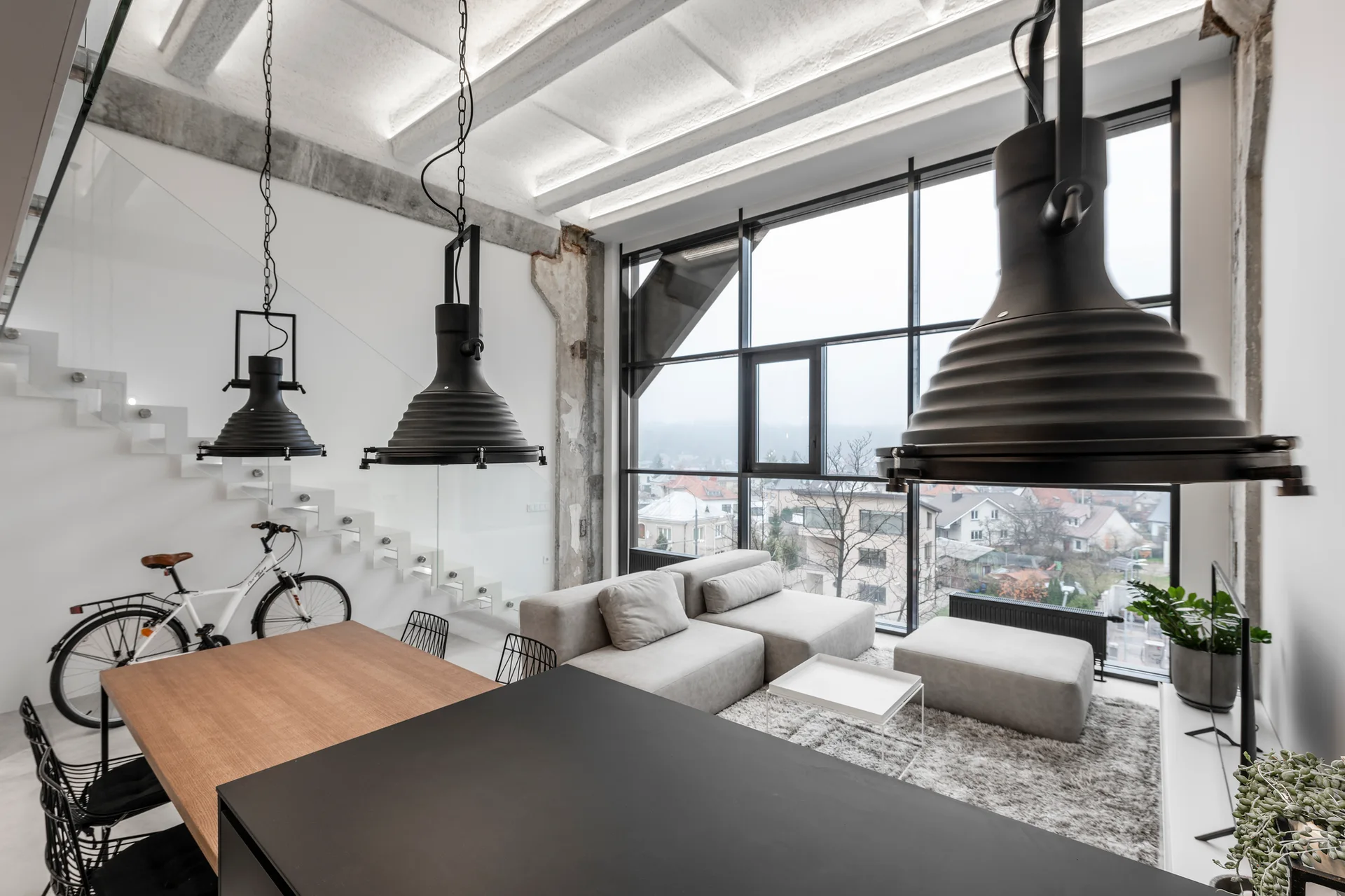 A modern industrial loft featuring a modular sofa, large black pendant lights, a floating staircase with glass railing, and a tall grid window.