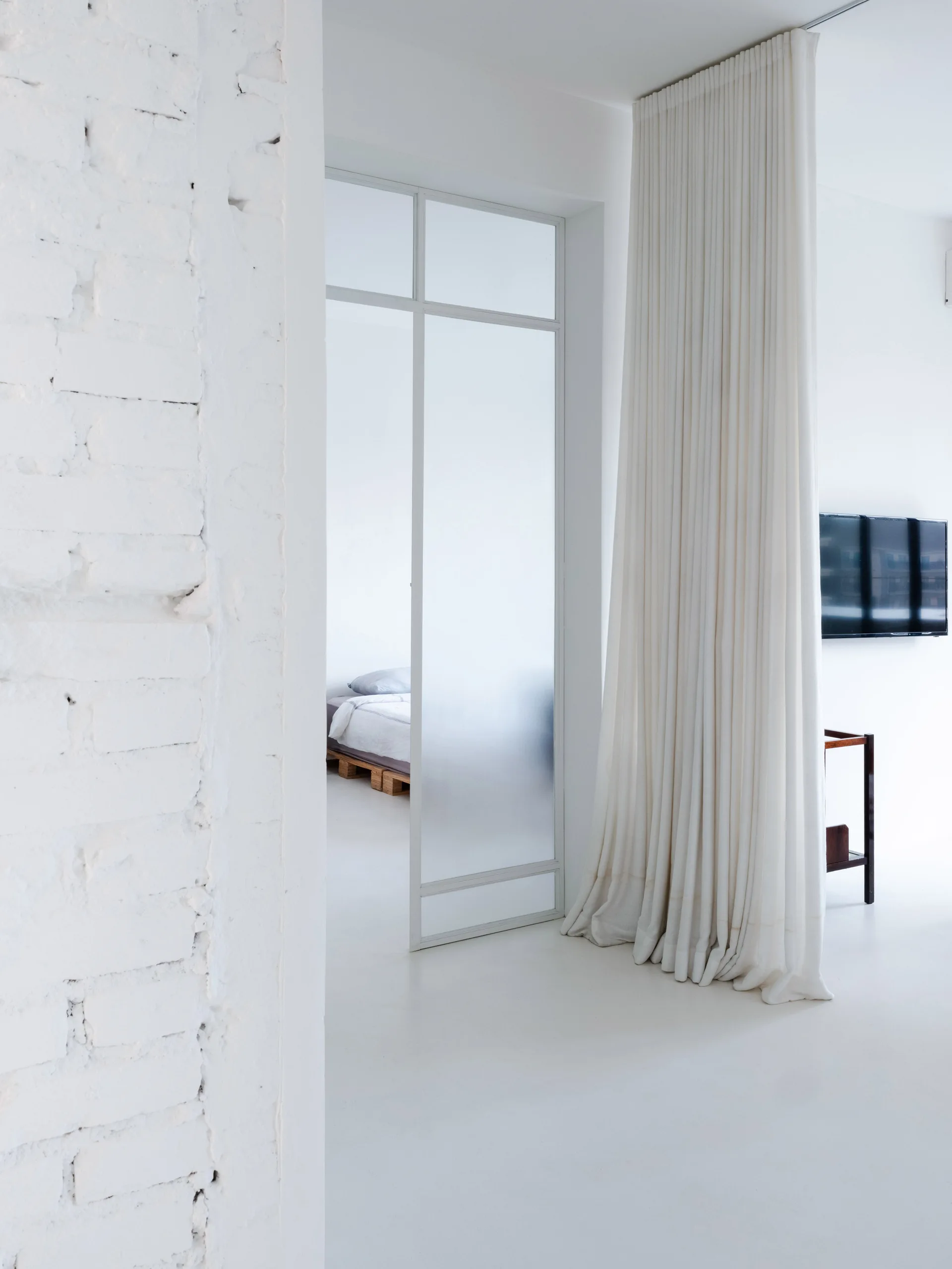 A white minimalist interior featuring an exposed white brick wall, a glass-paneled partition door, and a tall pleated white curtain.
