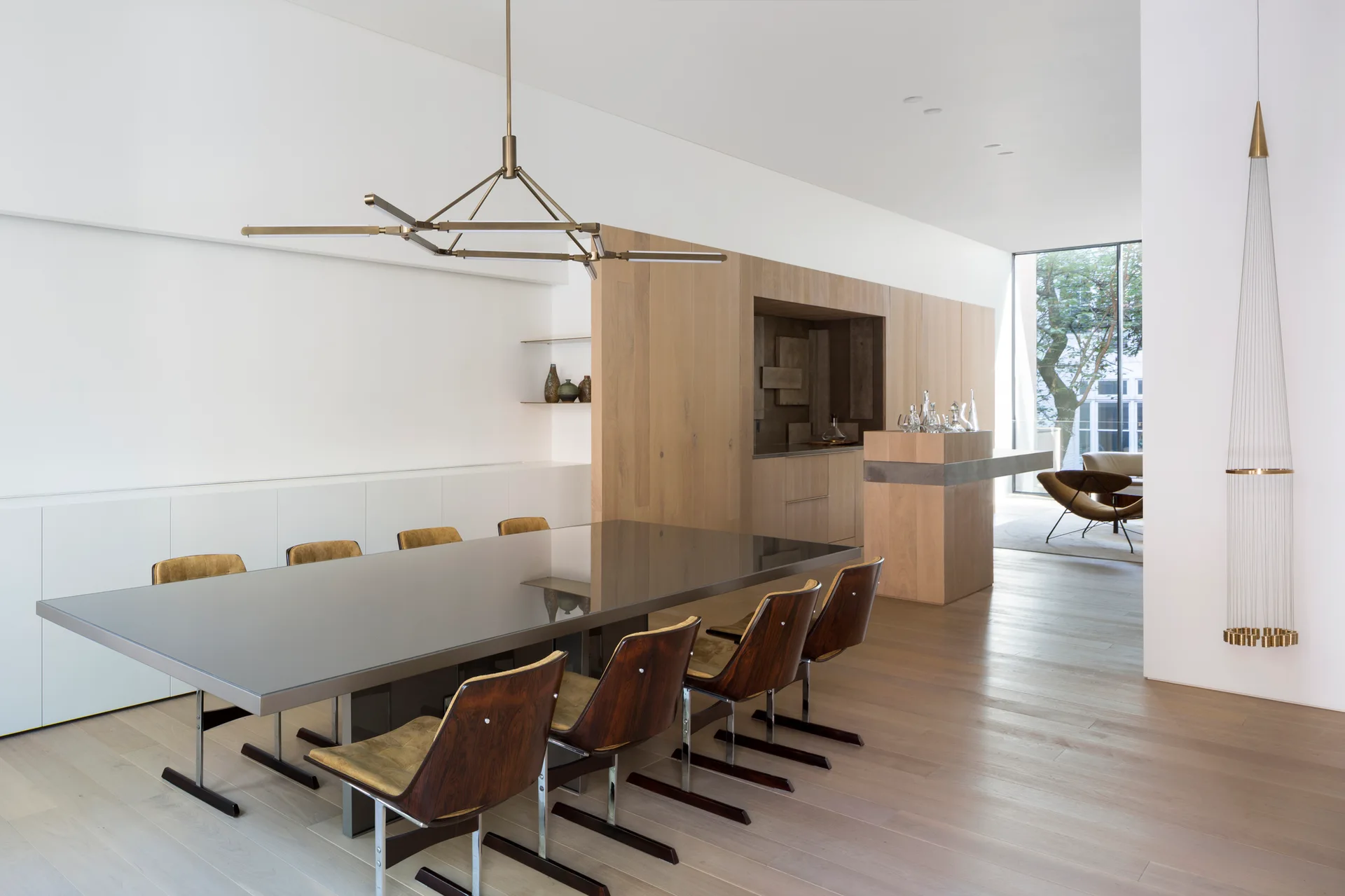 A modern dining room features a dark reflective table surrounded by mid-century wood chairs under a geometric brass pendant light.