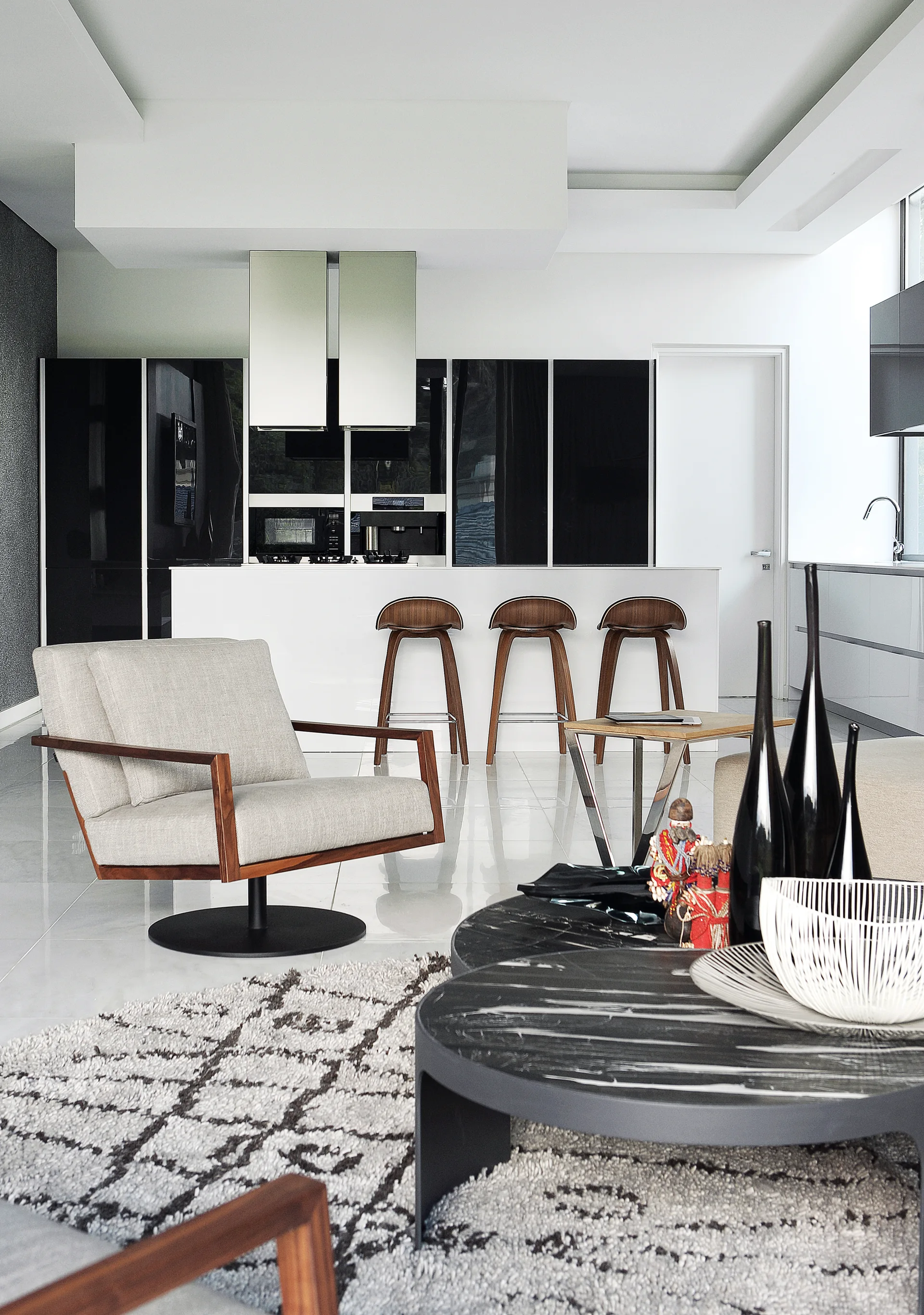 A modern open-concept space featuring a living area with a grey armchair and black coffee table next to a white kitchen island with three wooden stools.