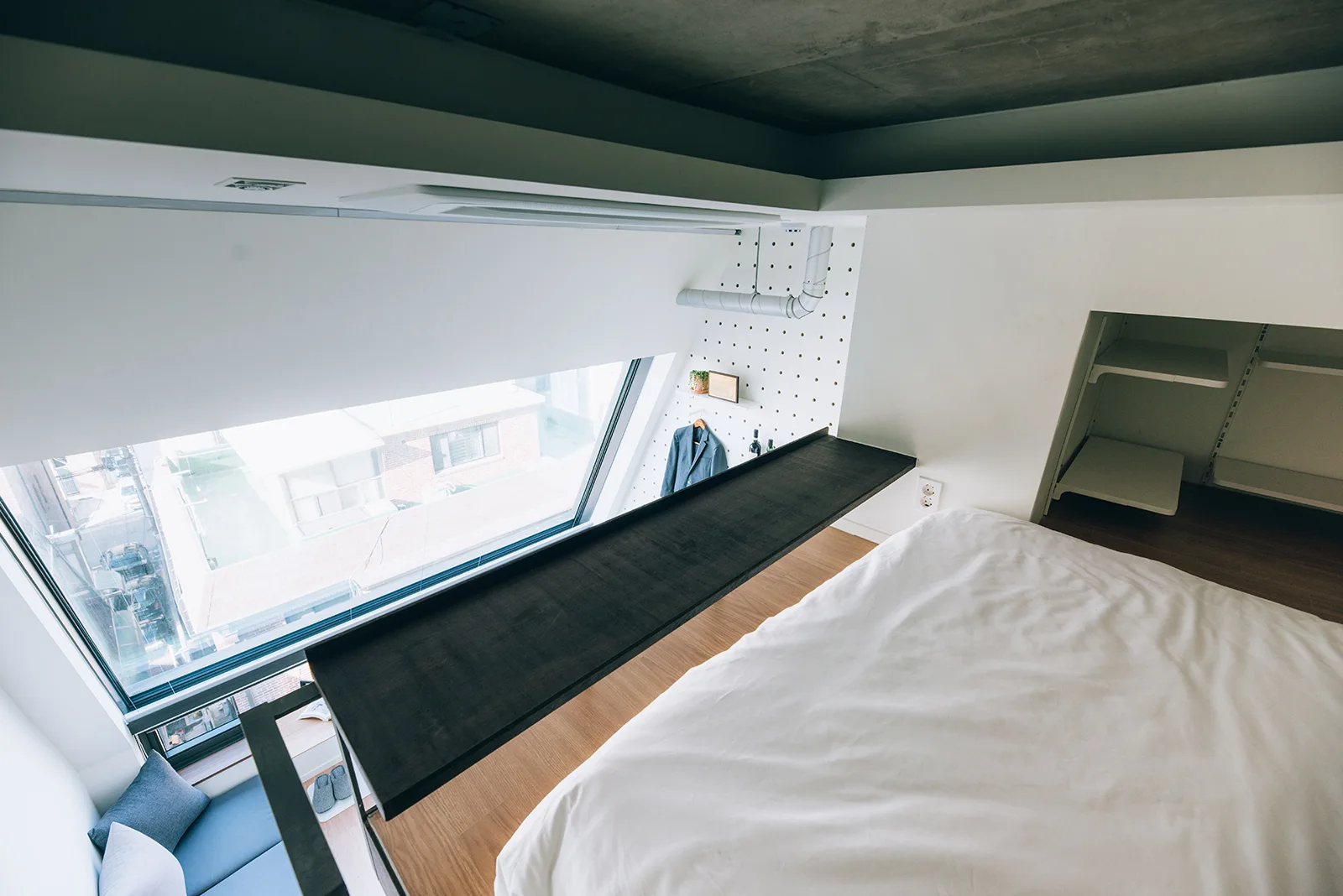 A loft bedroom featuring an elevated bed with white bedding, a sloped window, a dark wood ledge, and a white pegboard wall.