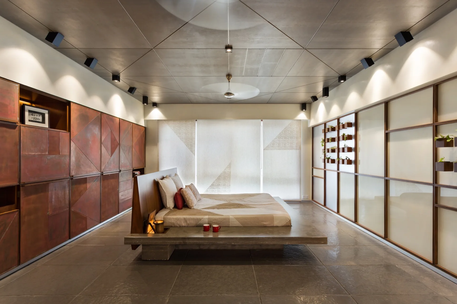 A minimalist bedroom features a low concrete platform bed, large rust-colored geometric wall cabinets, and a gridded concrete ceiling.