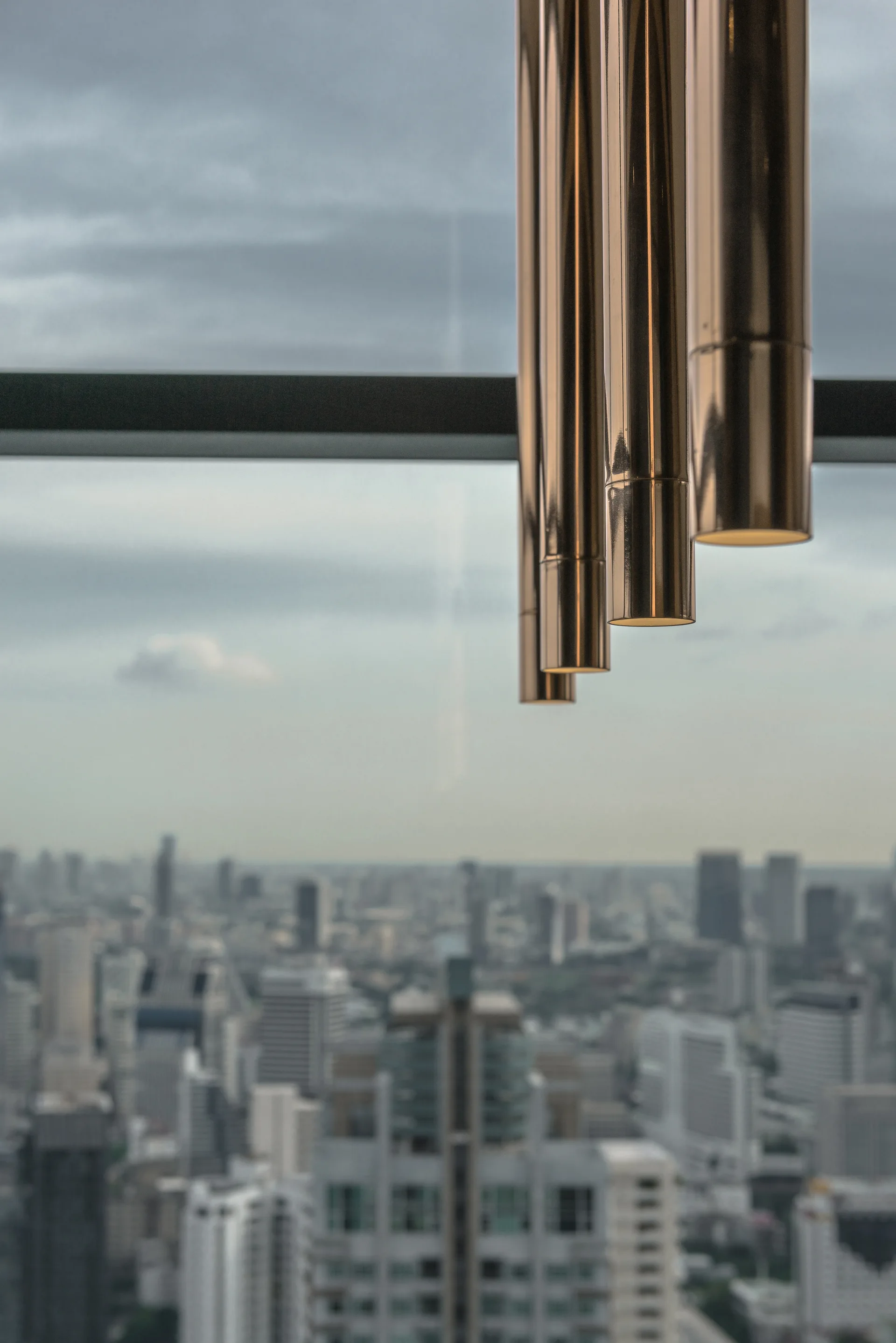 A cluster of polished bronze cylindrical pendant lights hangs in front of a window overlooking a hazy city skyline.