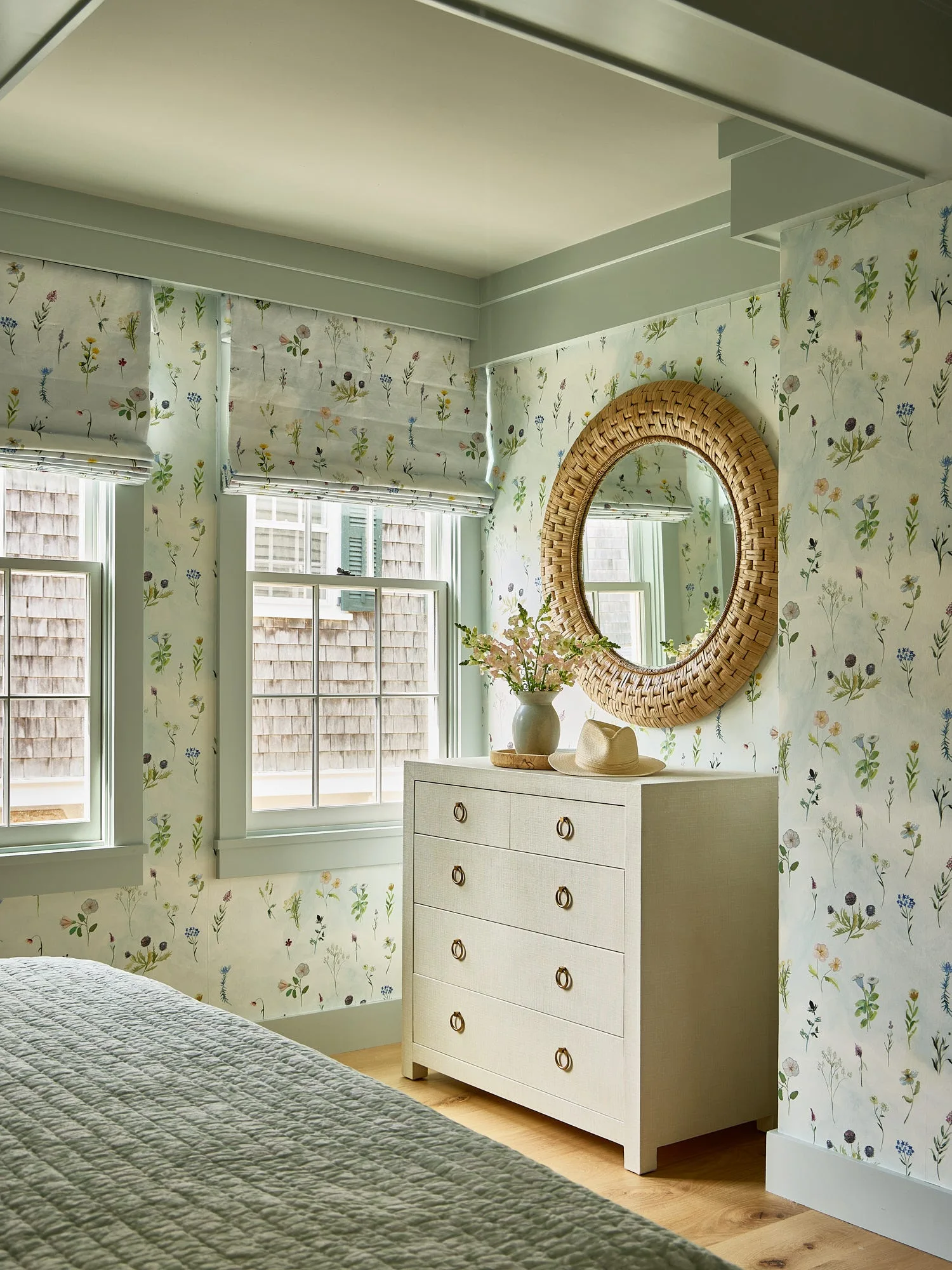 A cottage bedroom with botanical floral wallpaper, sage green window trim, a textured cream dresser, and a round woven mirror.