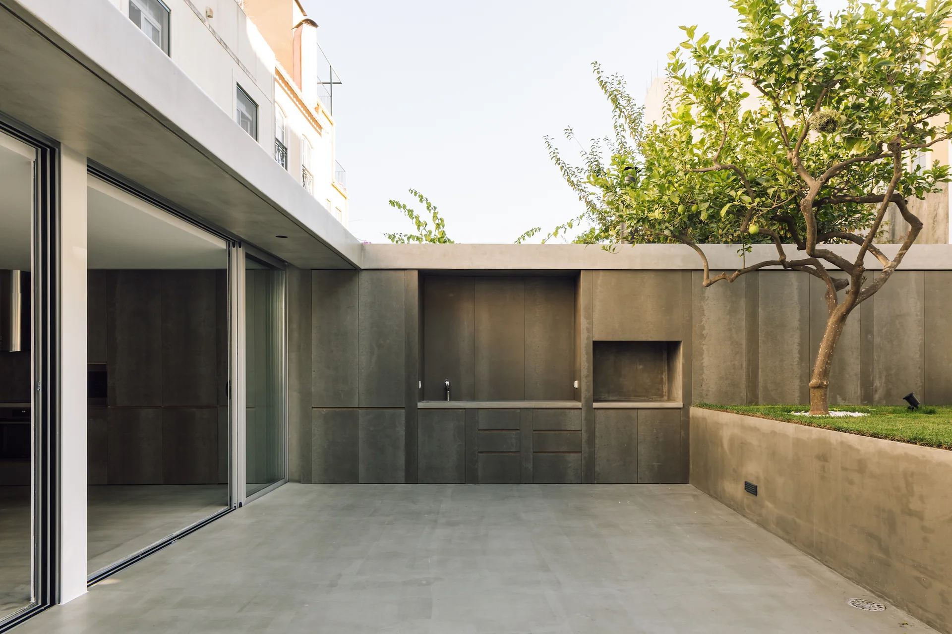 A minimalist concrete patio features a built-in grey kitchen station, sliding glass doors, and a single tree in a raised planter bed.