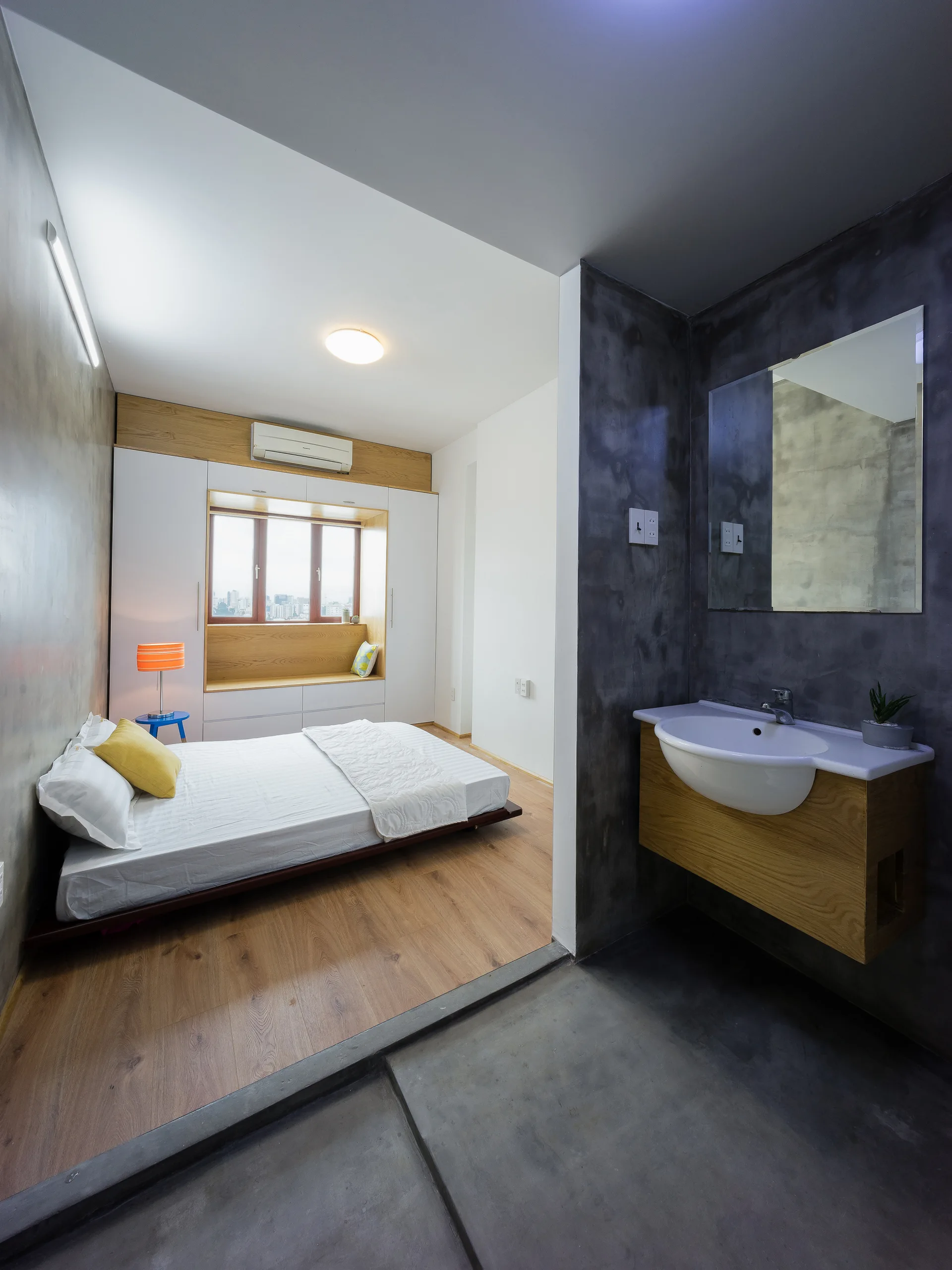 A modern bedroom with a low profile bed and window seat sits adjacent to a bathroom area with concrete walls and a wooden sink vanity.