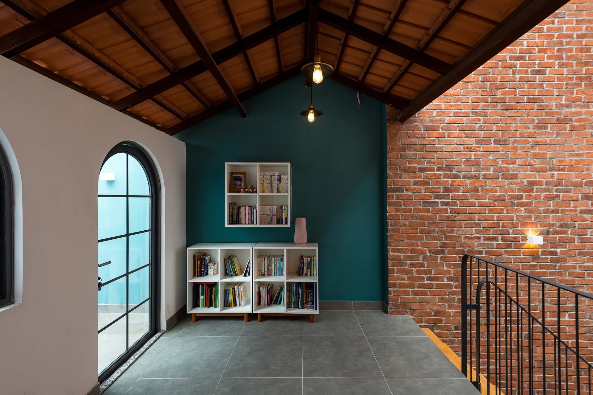A staircase landing features a white cubby bookshelf against a teal wall, alongside an exposed brick wall, arched window, and vaulted wood ceiling.