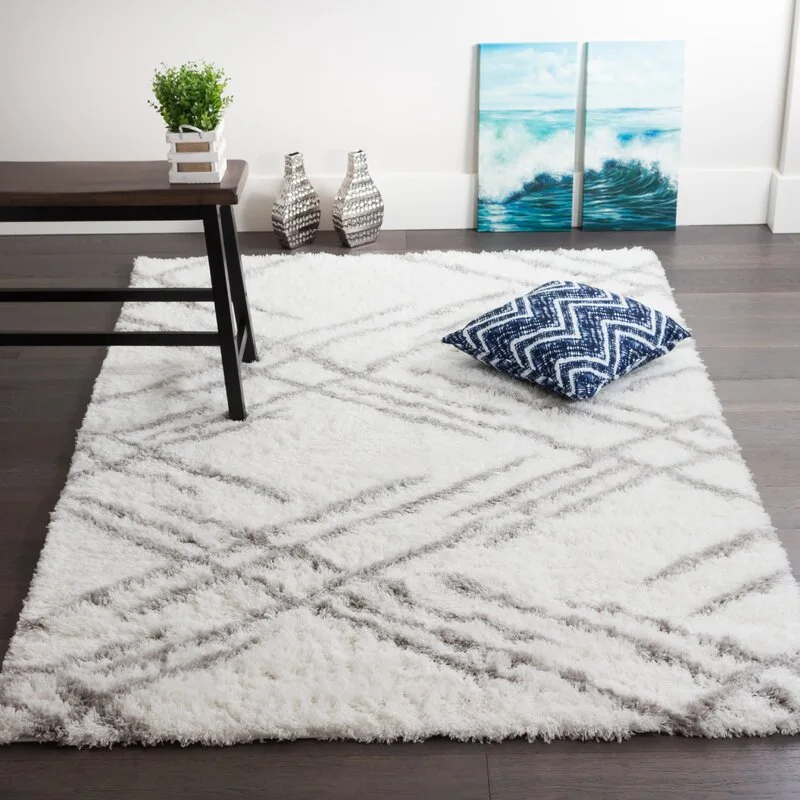 A plush white geometric shag rug rests on a dark wood floor alongside a blue patterned pillow, two silver vases, and coastal wall art.