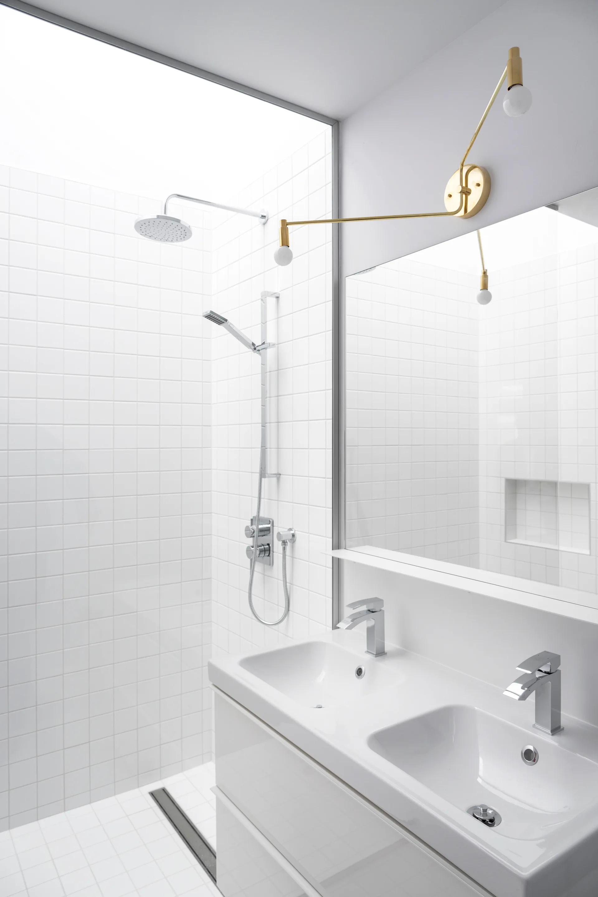 A modern white tiled bathroom featuring a double sink vanity, chrome fixtures, a large mirror, and an articulated brass wall light.