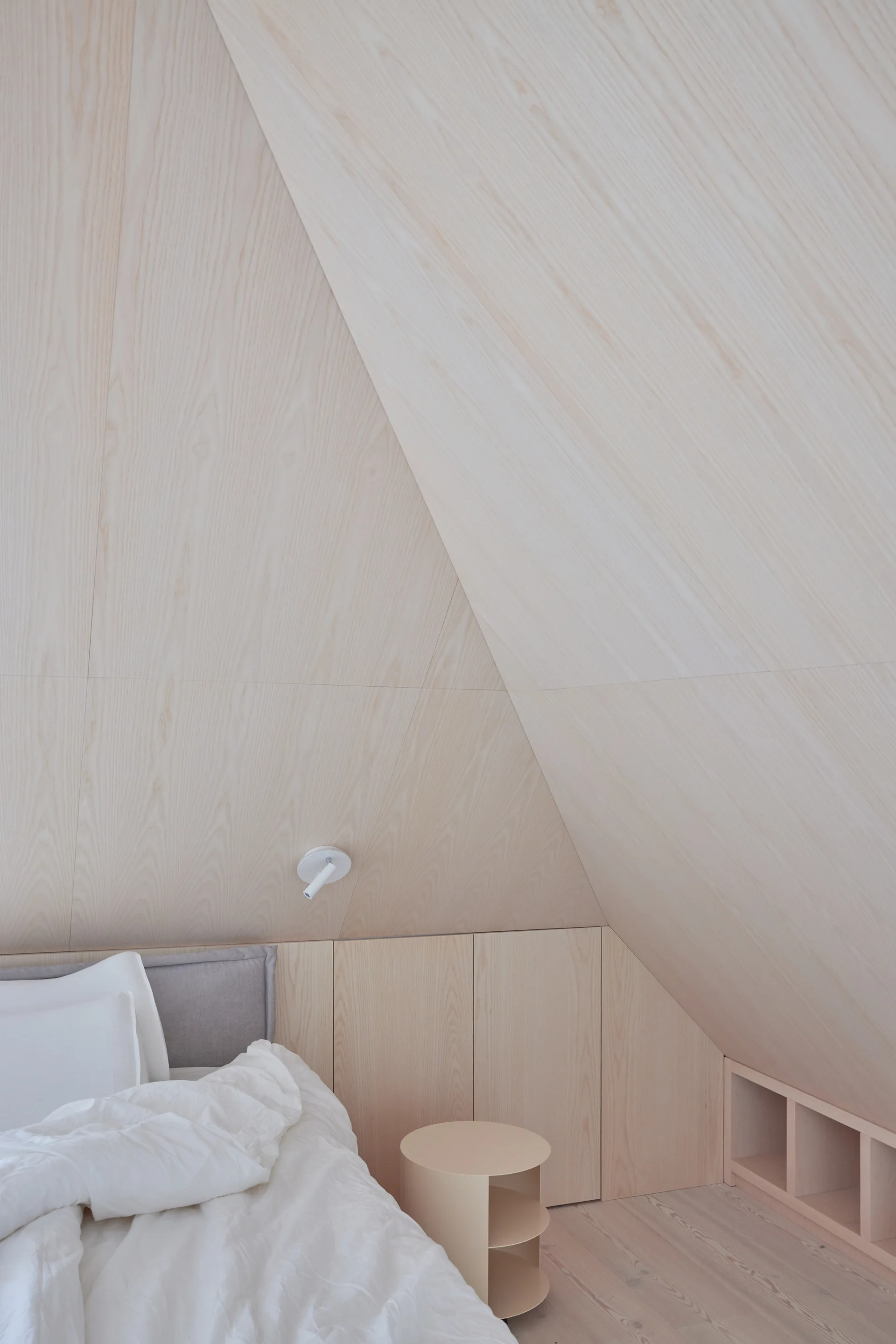 A minimalist attic bedroom features light wood-paneled walls and ceiling, built-in cabinets, and a bed with white sheets.