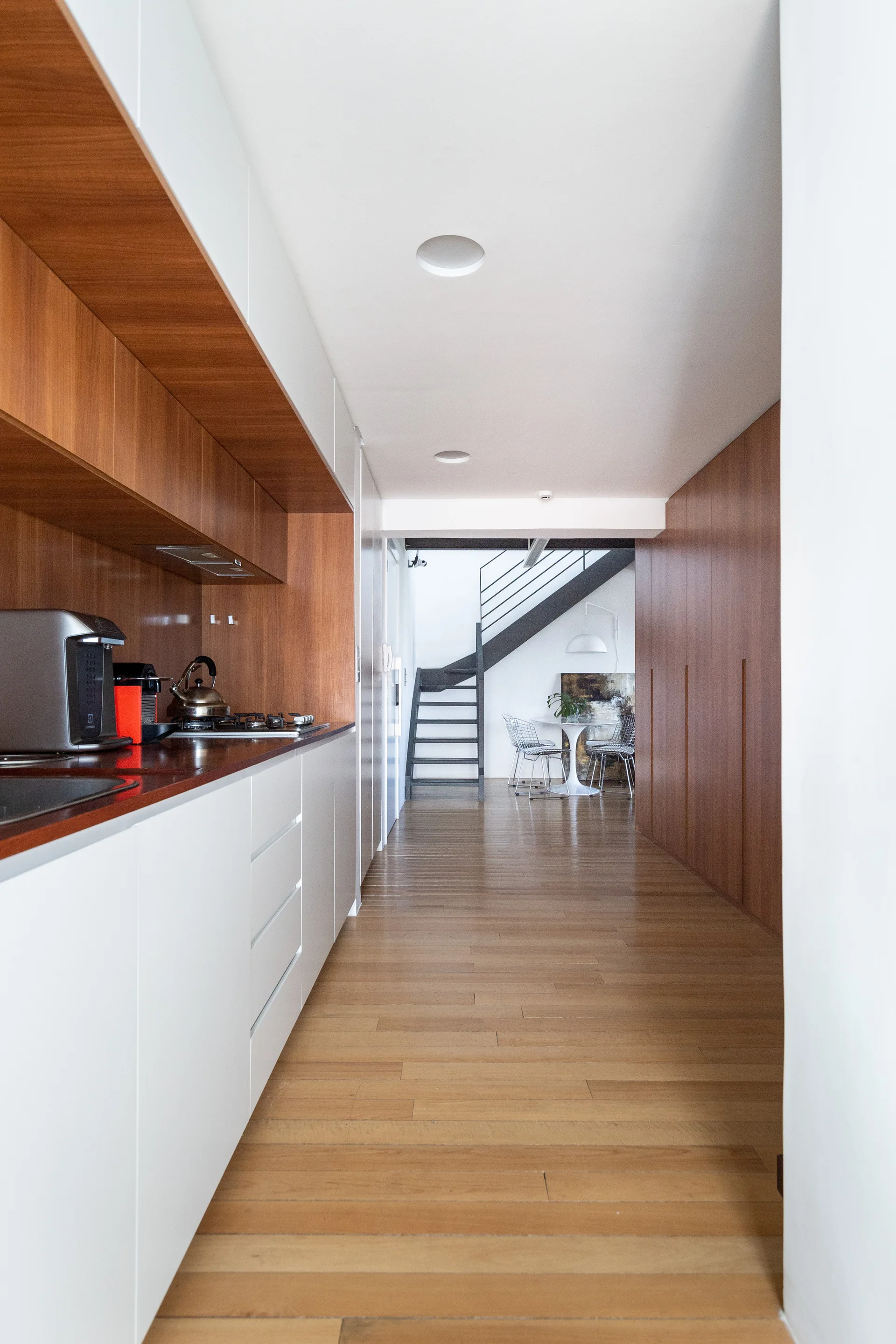 A long galley kitchen features white lower cabinets and warm wood-paneled walls leading to a dining area with a metal staircase.