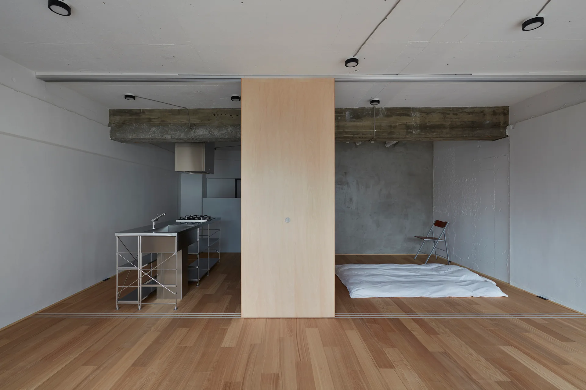 A minimalist studio apartment with a wood sliding room divider, a stainless steel kitchen island, and concrete ceilings.