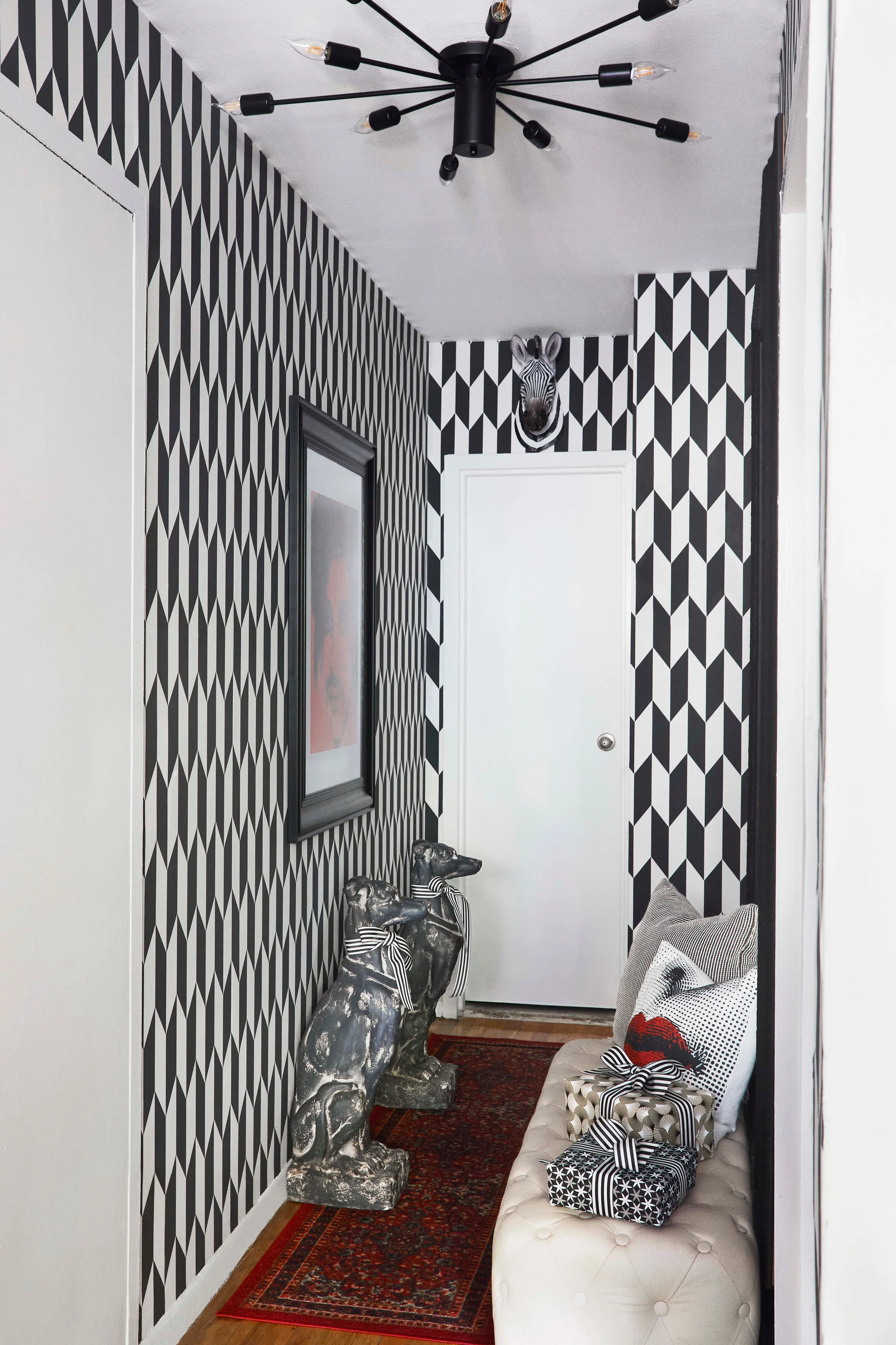 A narrow hallway featuring black and white geometric wallpaper, a tufted cream bench with decorative pillows, and dark dog statues on a red runner.